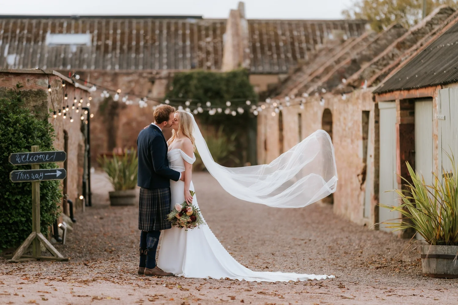 The couple kiss beside the stone buildings at The Cow Shed Crail, with the bride’s veil billowing behind her.