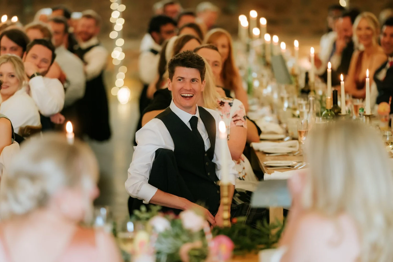 The groom turns towards the speaker while seated at the long candlelit wedding table.