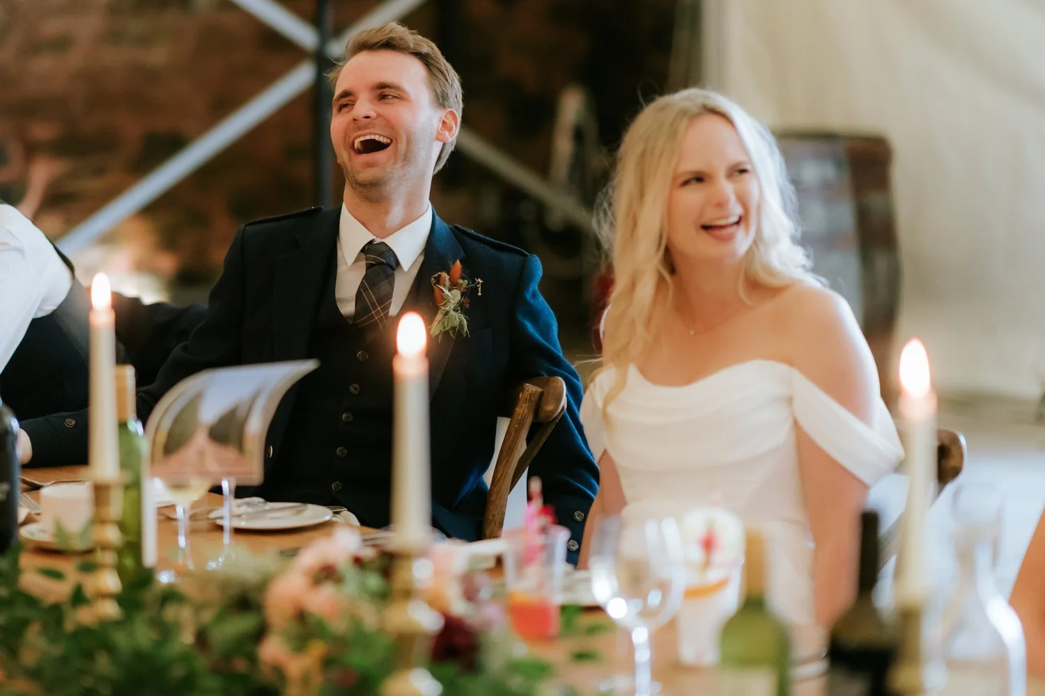 The couple laugh at the top table, surrounded by candles, glassware and flowers.