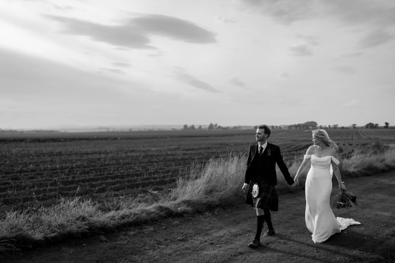 Black-and-white photograph of the couple walking beside open farmland beneath a clouded sky.