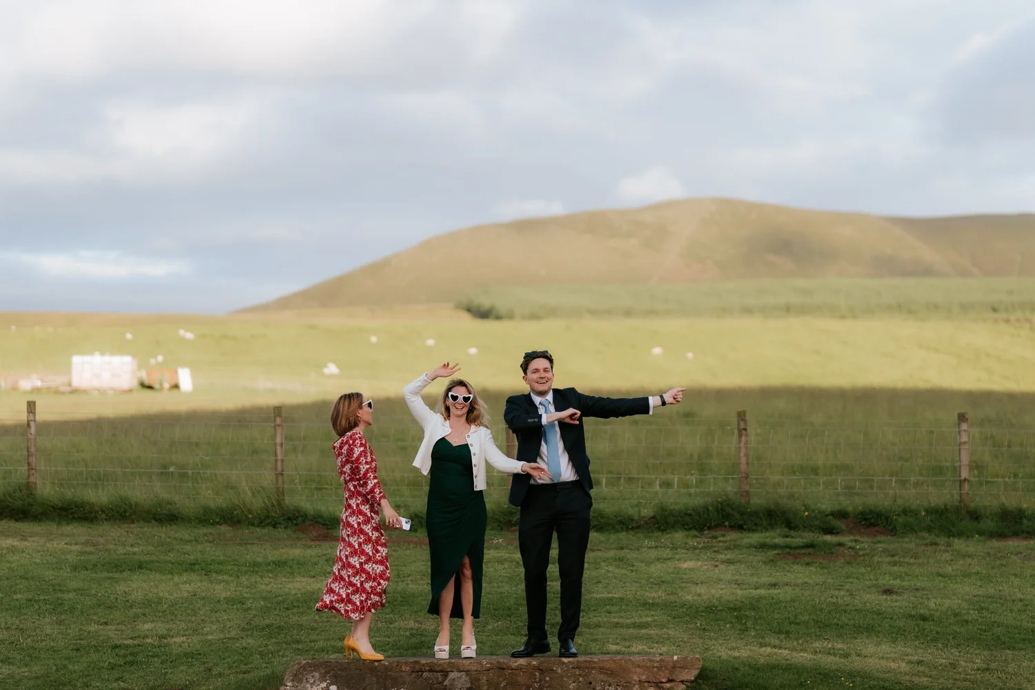 Three guests posing with their arms outstretched on the lawn beneath the hills.