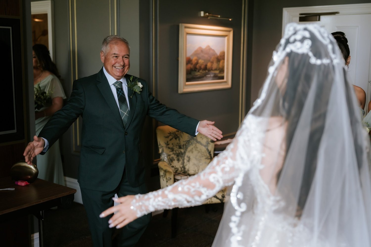 A smiling man opening his arms towards Cristina as she stands in her wedding dress and veil.
