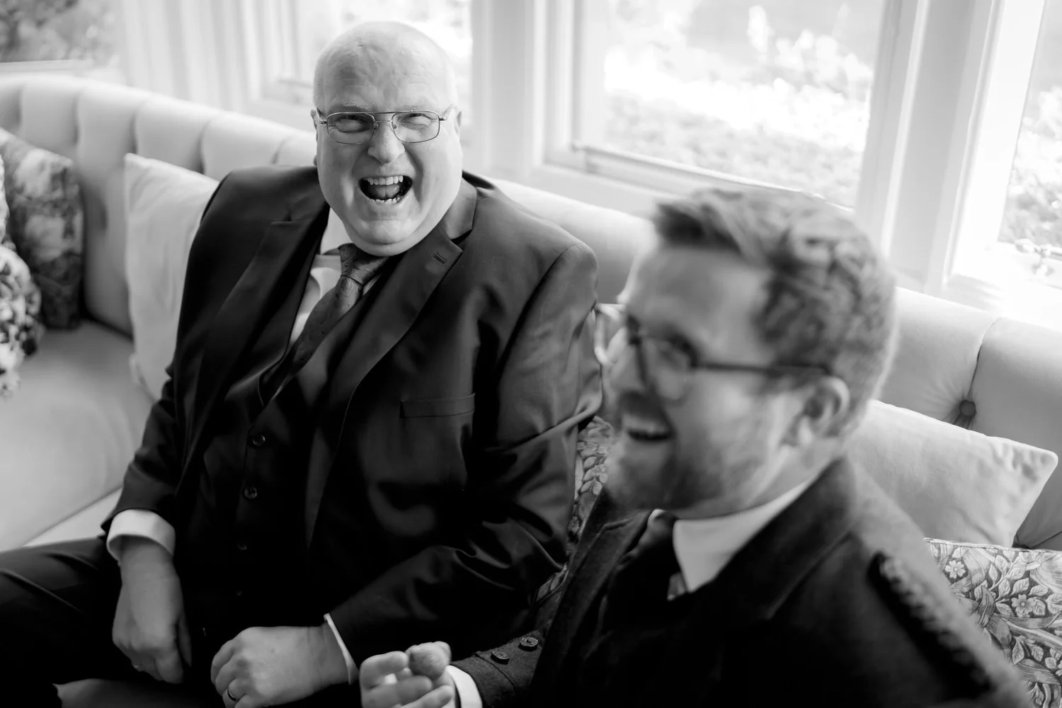Two wedding guests laughing together on a sofa, photographed in black and white.