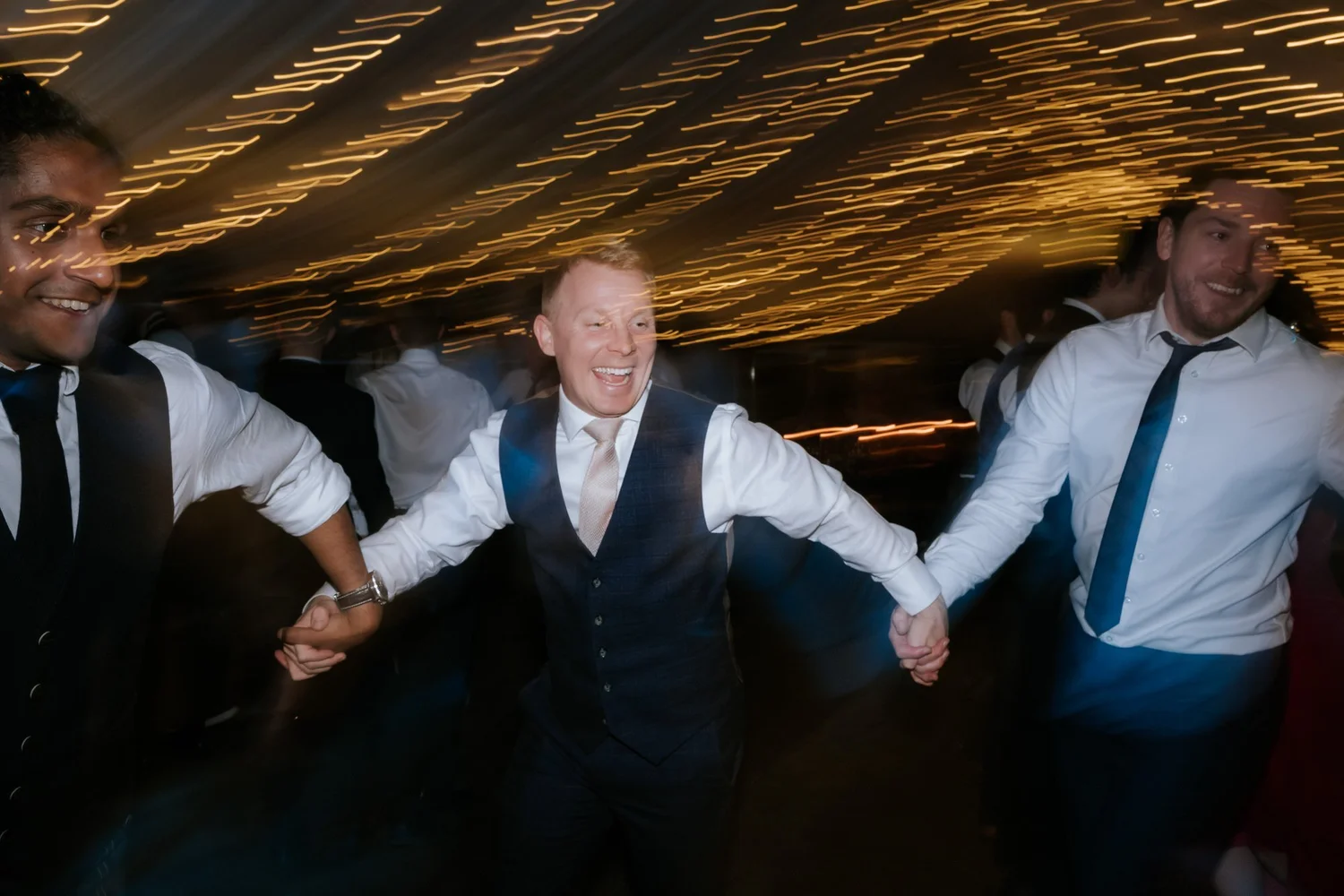 Guests hold hands during a lively ceilidh beneath the reception marquee’s canopy of lights.