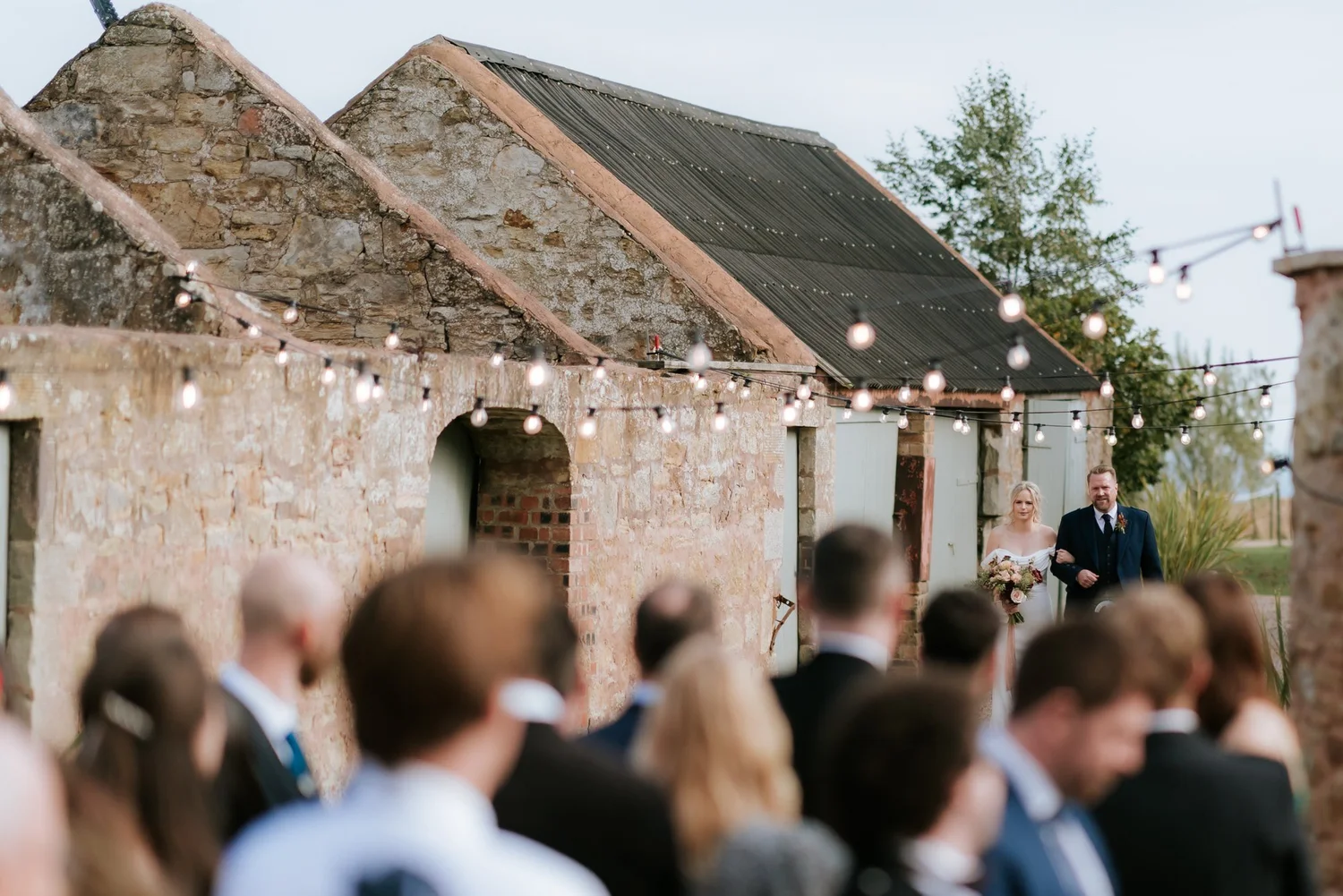 A view past seated wedding guests towards a man standing beneath festoon lights beside the stone building.