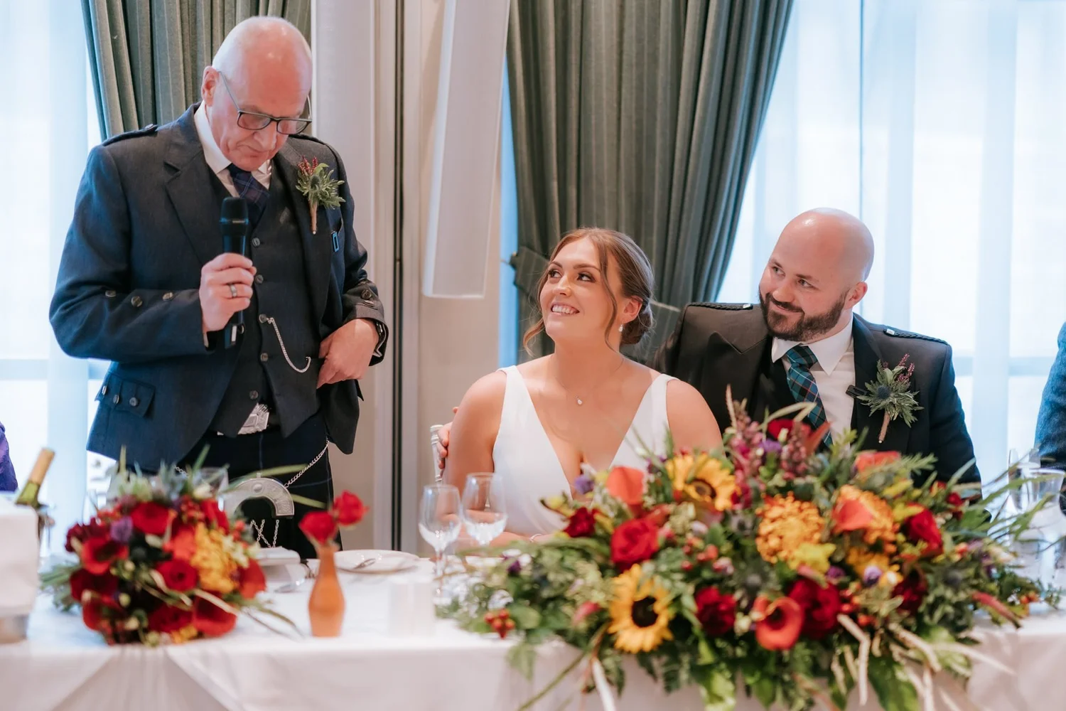 Kendall and Ewen looking up at her dad as he gives his wedding speech beside the autumn flowers.