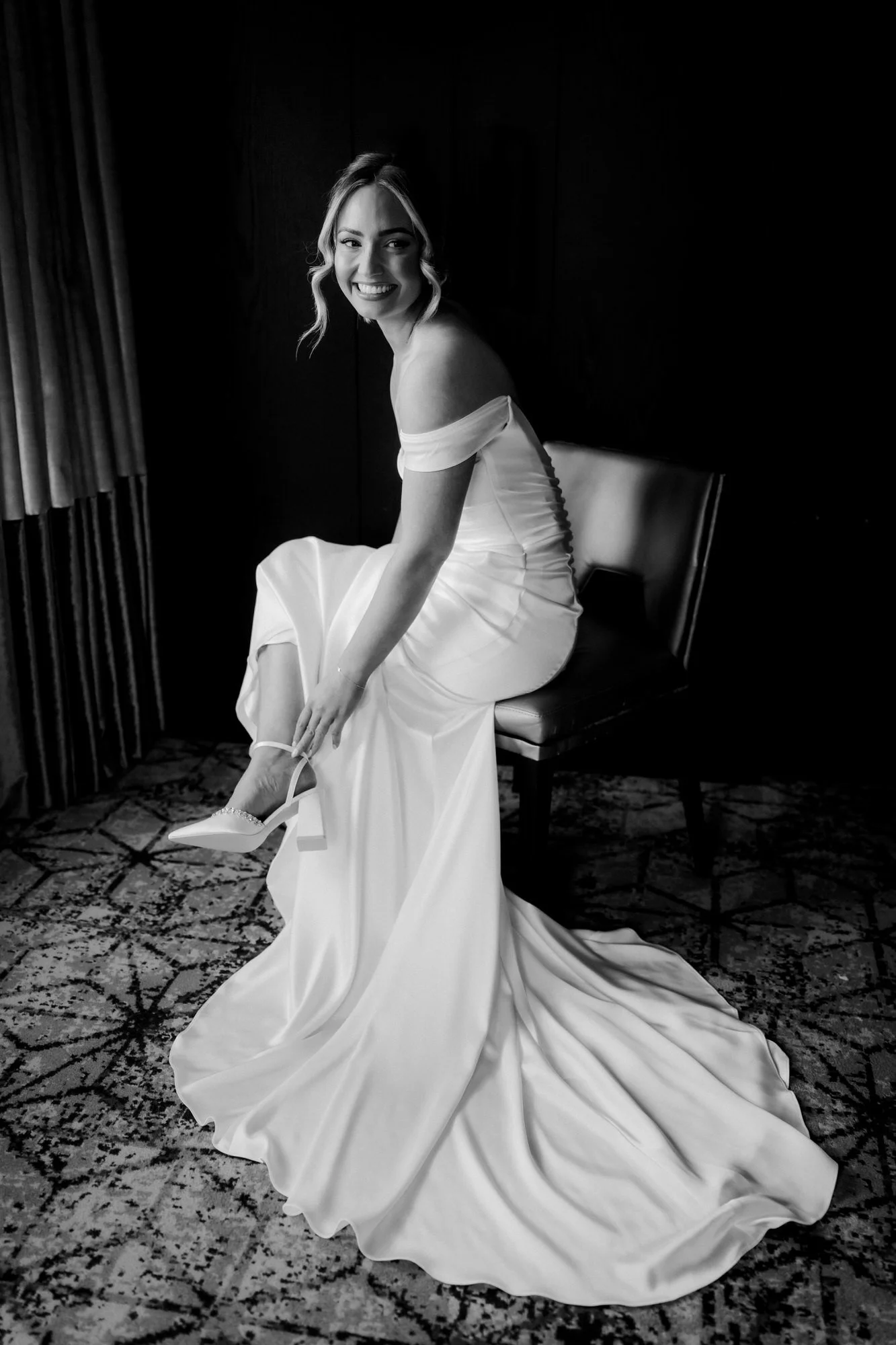 Black-and-white portrait of Elle lifting the hem of her wedding dress beside a window