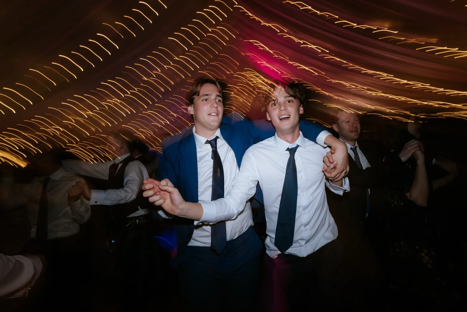 Two guests dance shoulder to shoulder on the crowded dance floor, with streaks of warm light overhead.