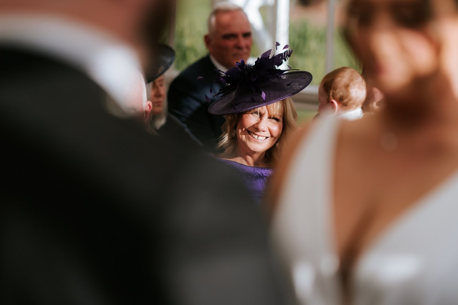 A guest in a purple hat smiling during the ceremony, framed between Kendall and Ewen.