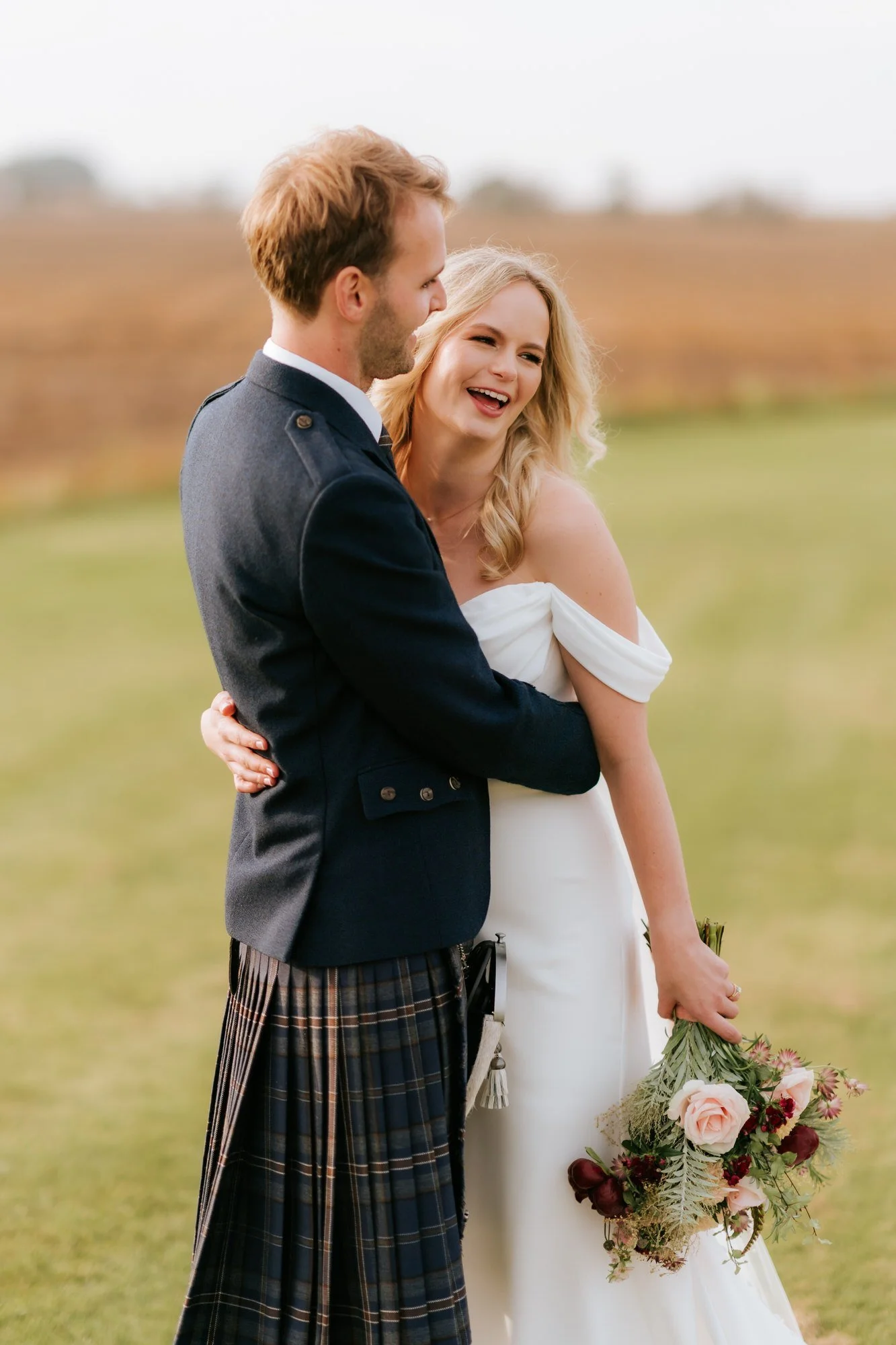 The bride laughs while the groom holds her close, with her bouquet at her side.