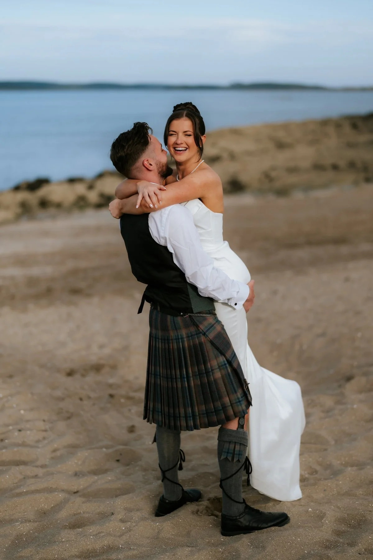 Lois laughing as Dougie lifts her into his arms on the beach by the water.