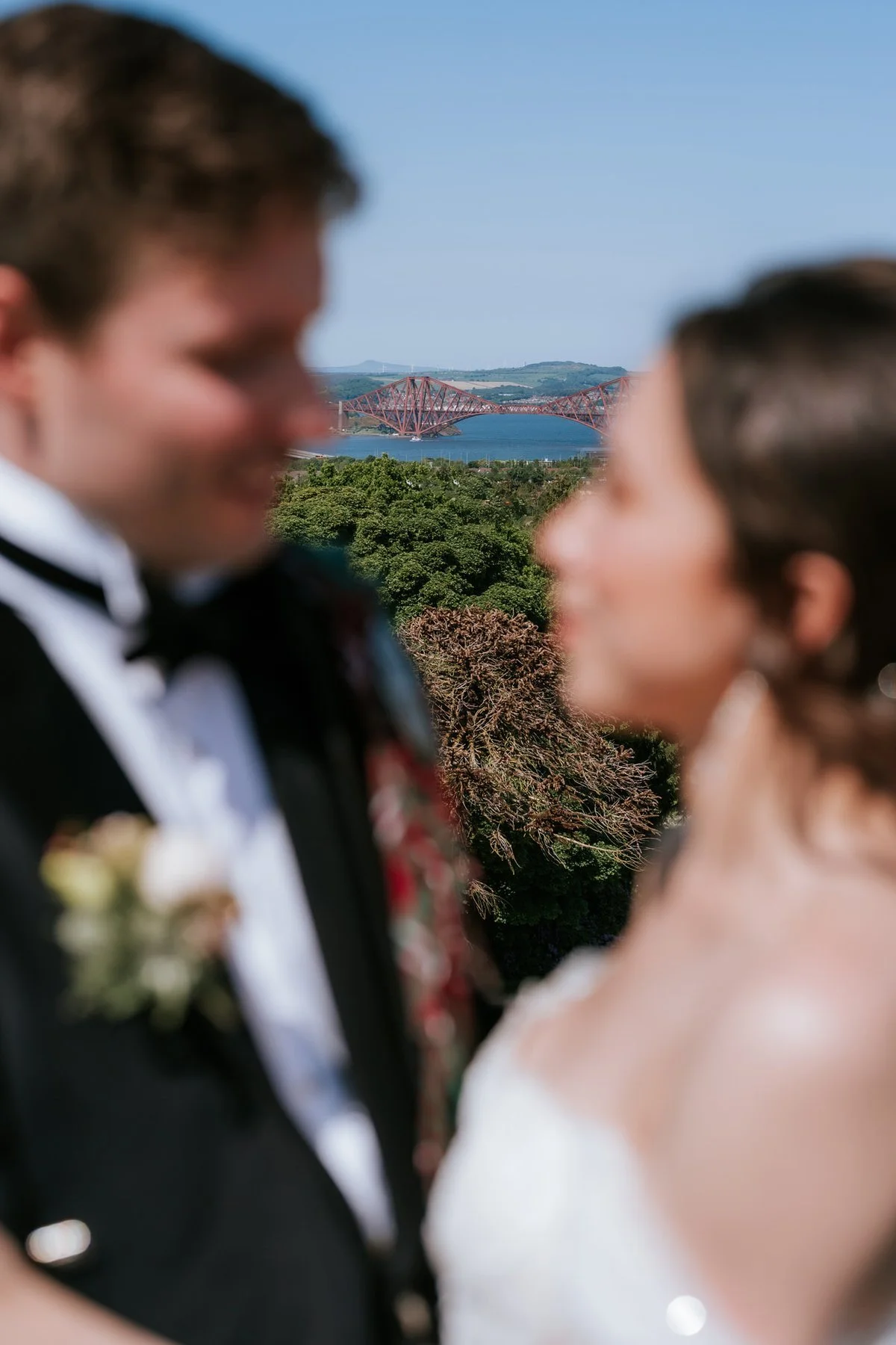 The Forth Bridge in focus beyond Kim and Kenneth, who are softly blurred in the foreground of this rooftop portrait.