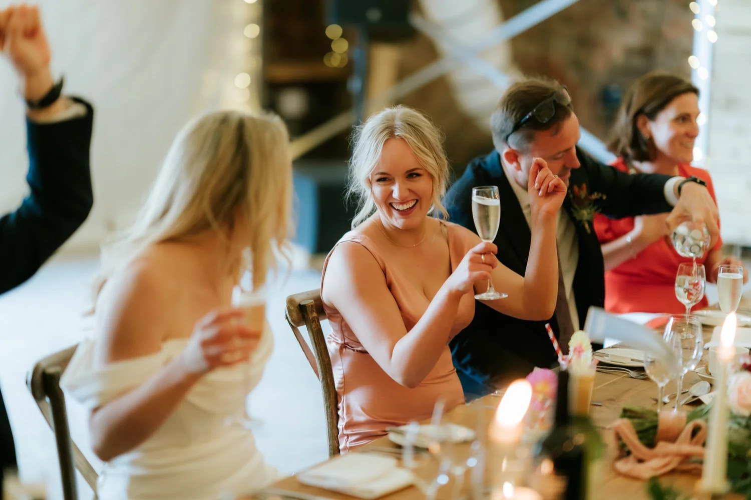 A guest holding a sparkling-wine glass laughs as another covers part of his face beside her.