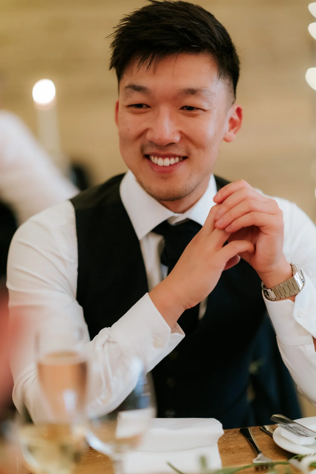 A guest in a white shirt and black waistcoat smiles with his hands clasped beside his face.