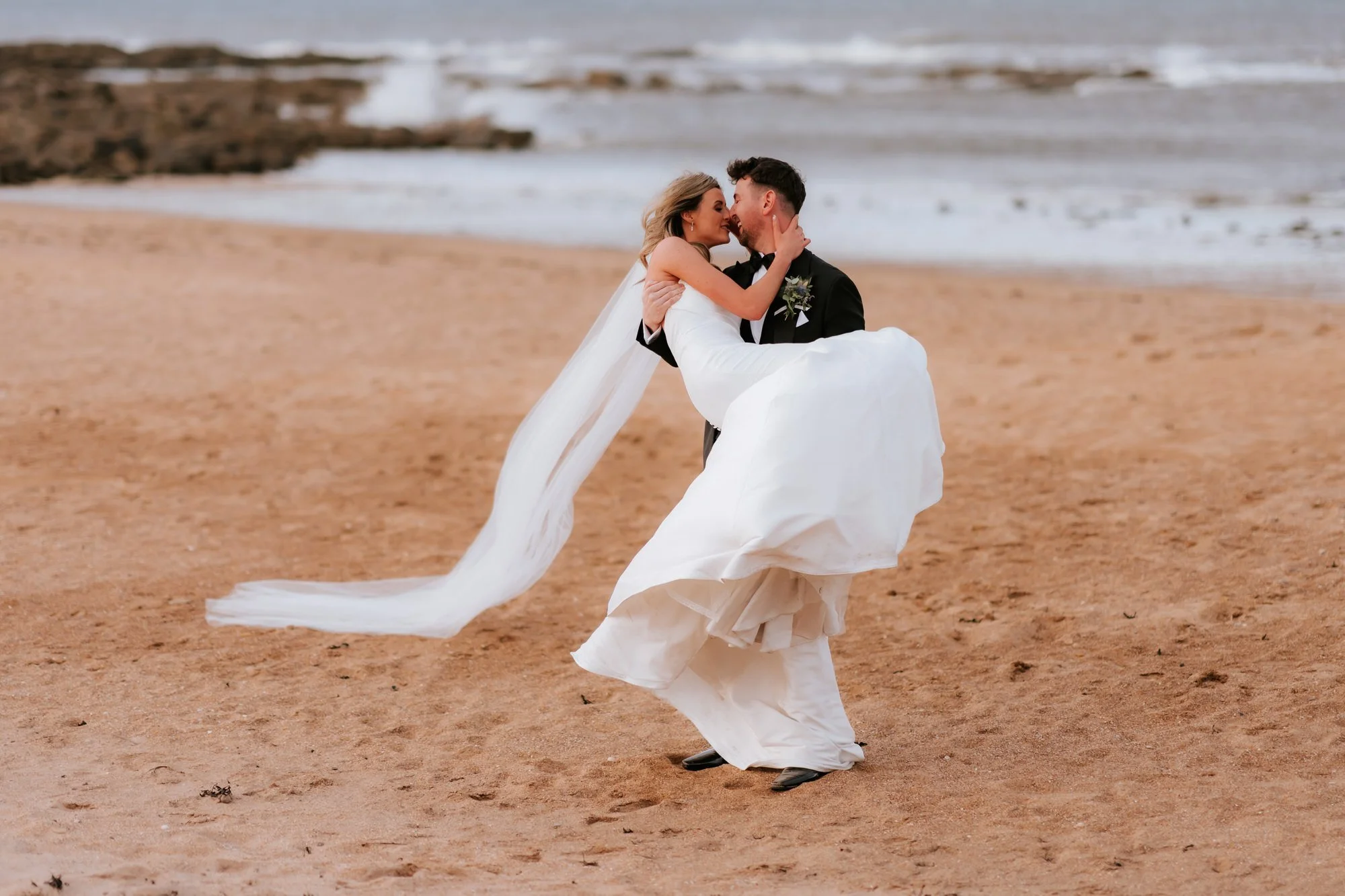 Jamie carrying Natalie as they kiss beside the sea