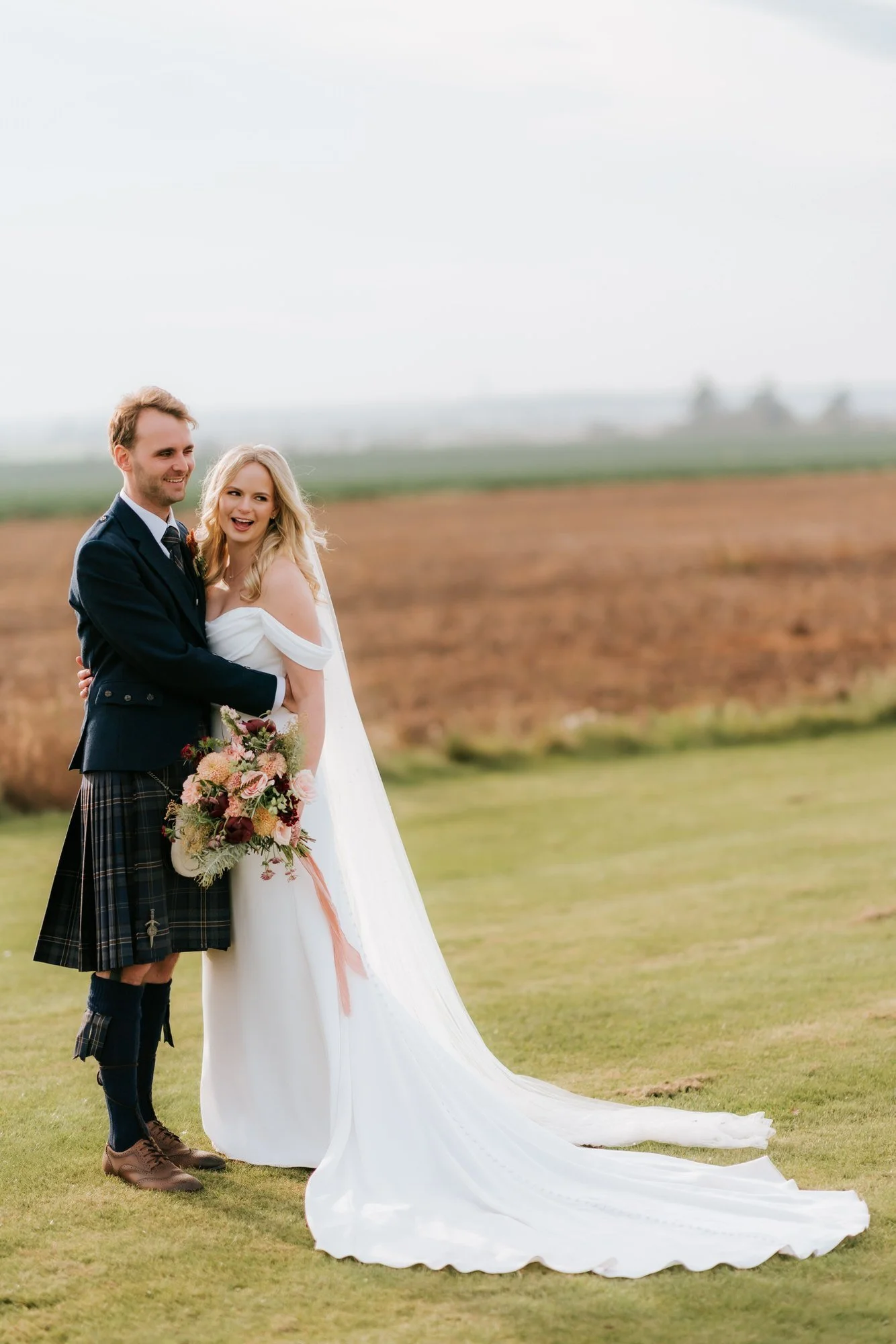 The couple stand together beside the fields, the bride holding a bouquet above her flowing train.