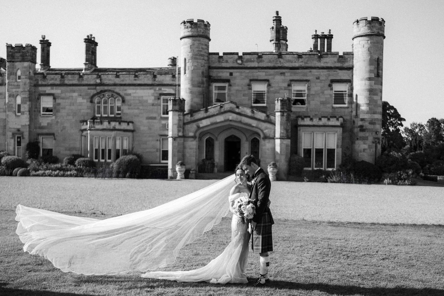 Black-and-white portrait of Kim and Kenneth kissing on the lawn, with her veil blowing out beside them and the castle behind.
