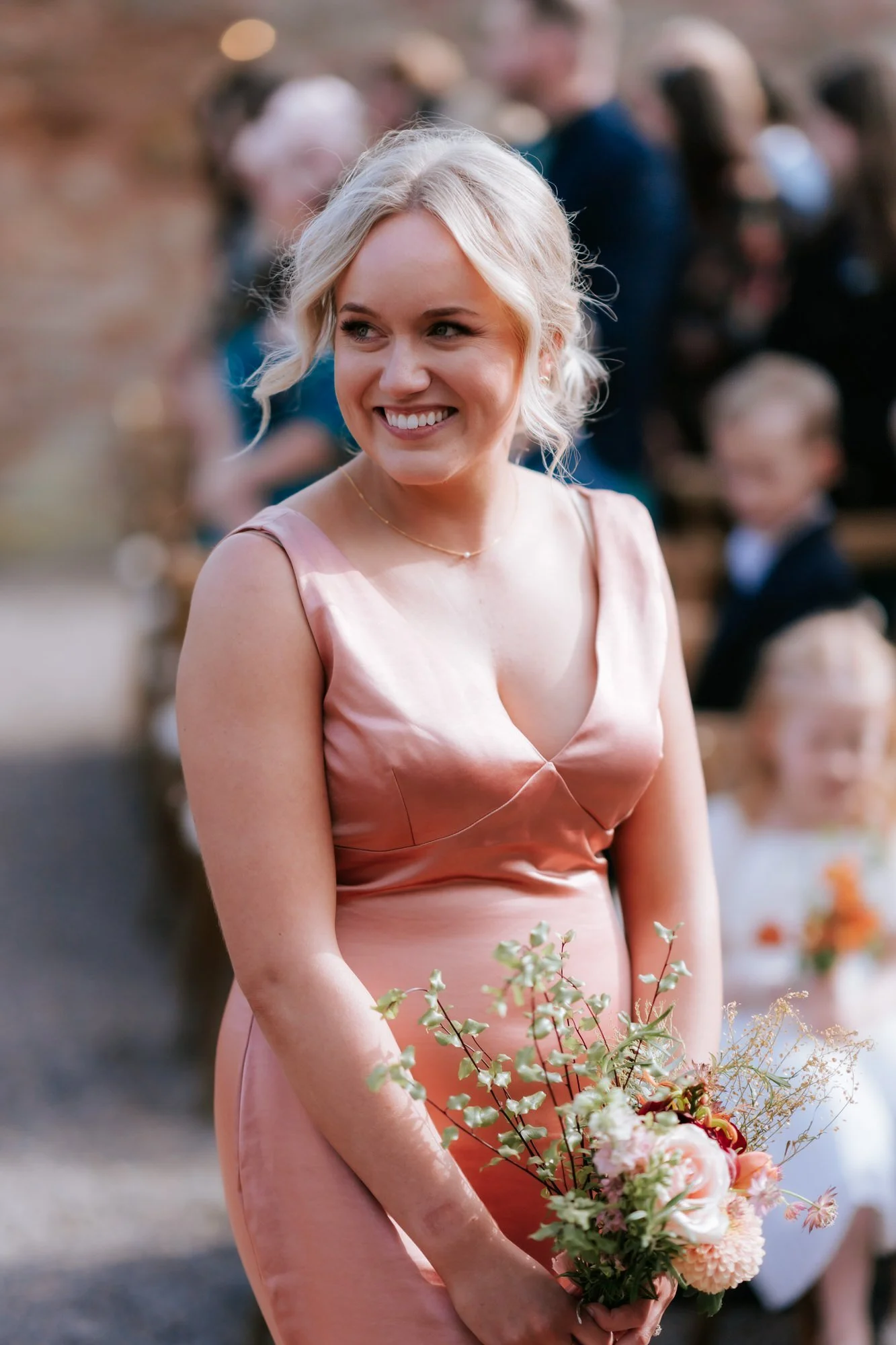 A bridesmaid in a pink dress smiles while holding her bouquet during the outdoor ceremony.