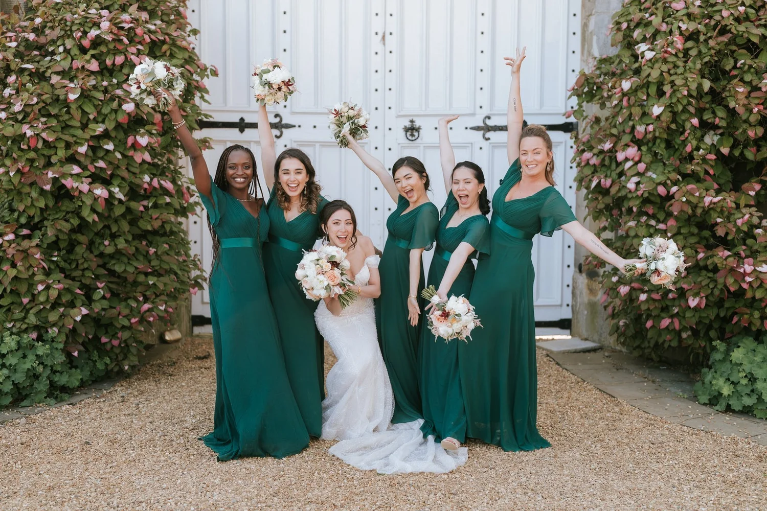 Kim and her bridesmaids raising their bouquets beside the flower-covered entrance at Dundas Castle.