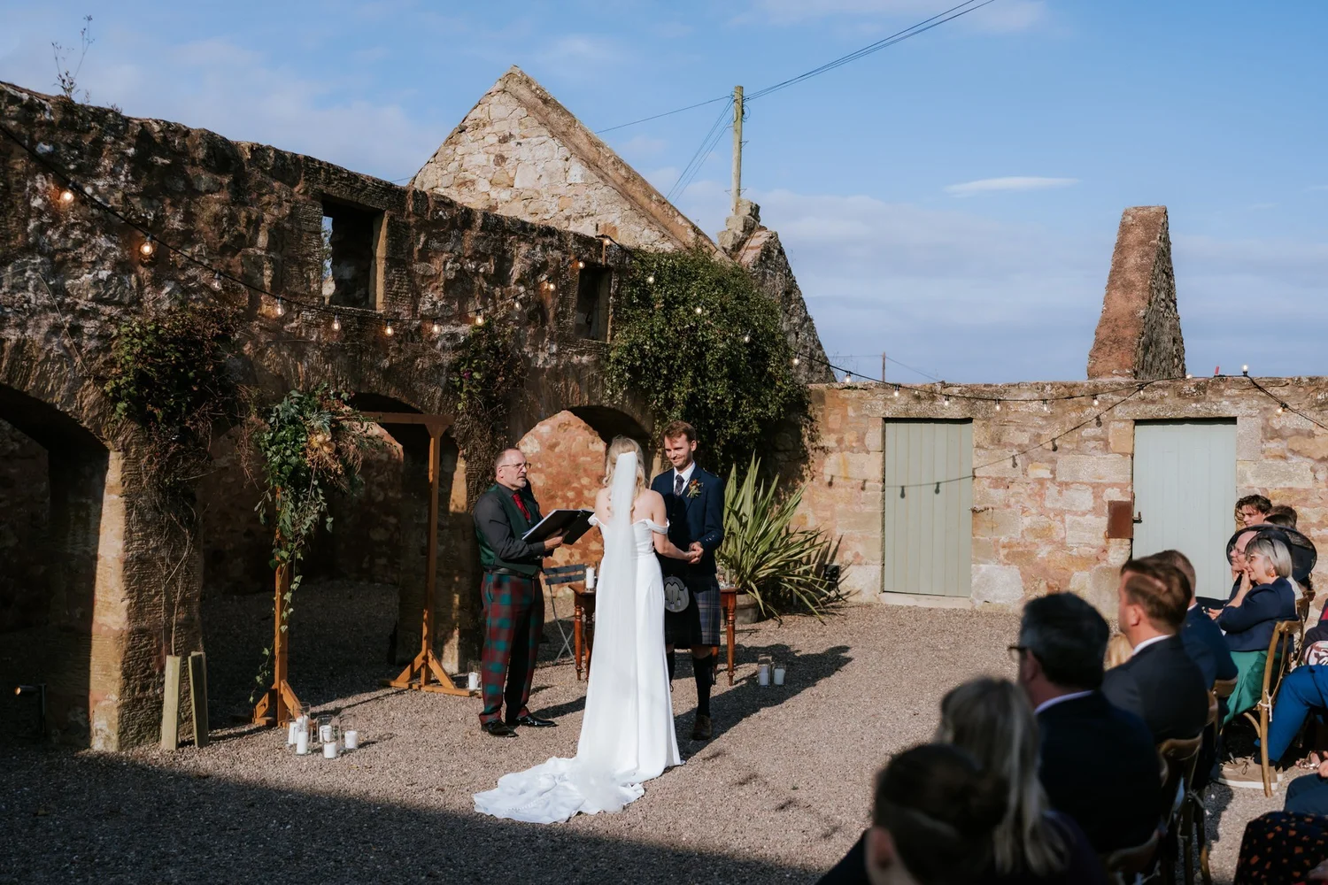 The outdoor wedding ceremony framed by the venue’s stone buildings, with guests seated in the sunshine.
