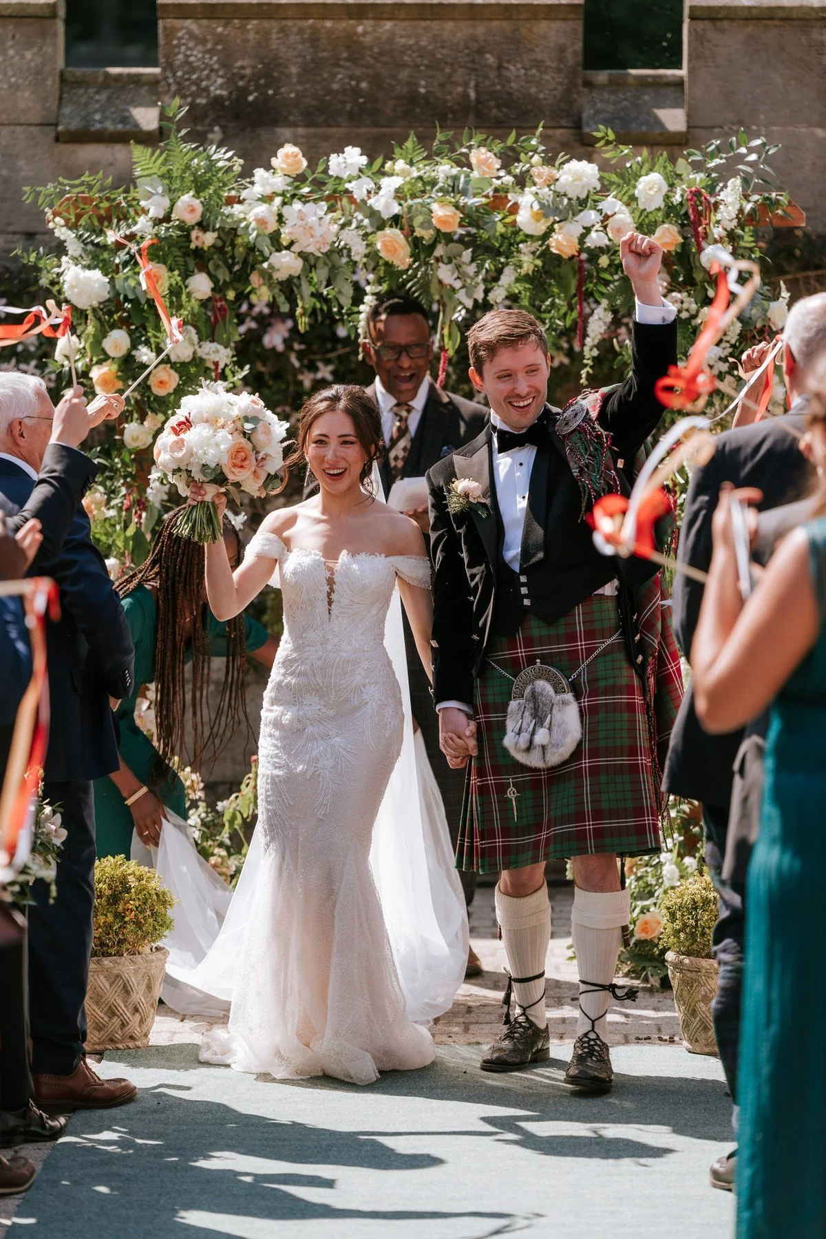 Kim and Kenneth walking back down the outdoor aisle with their arms raised as guests cheer.