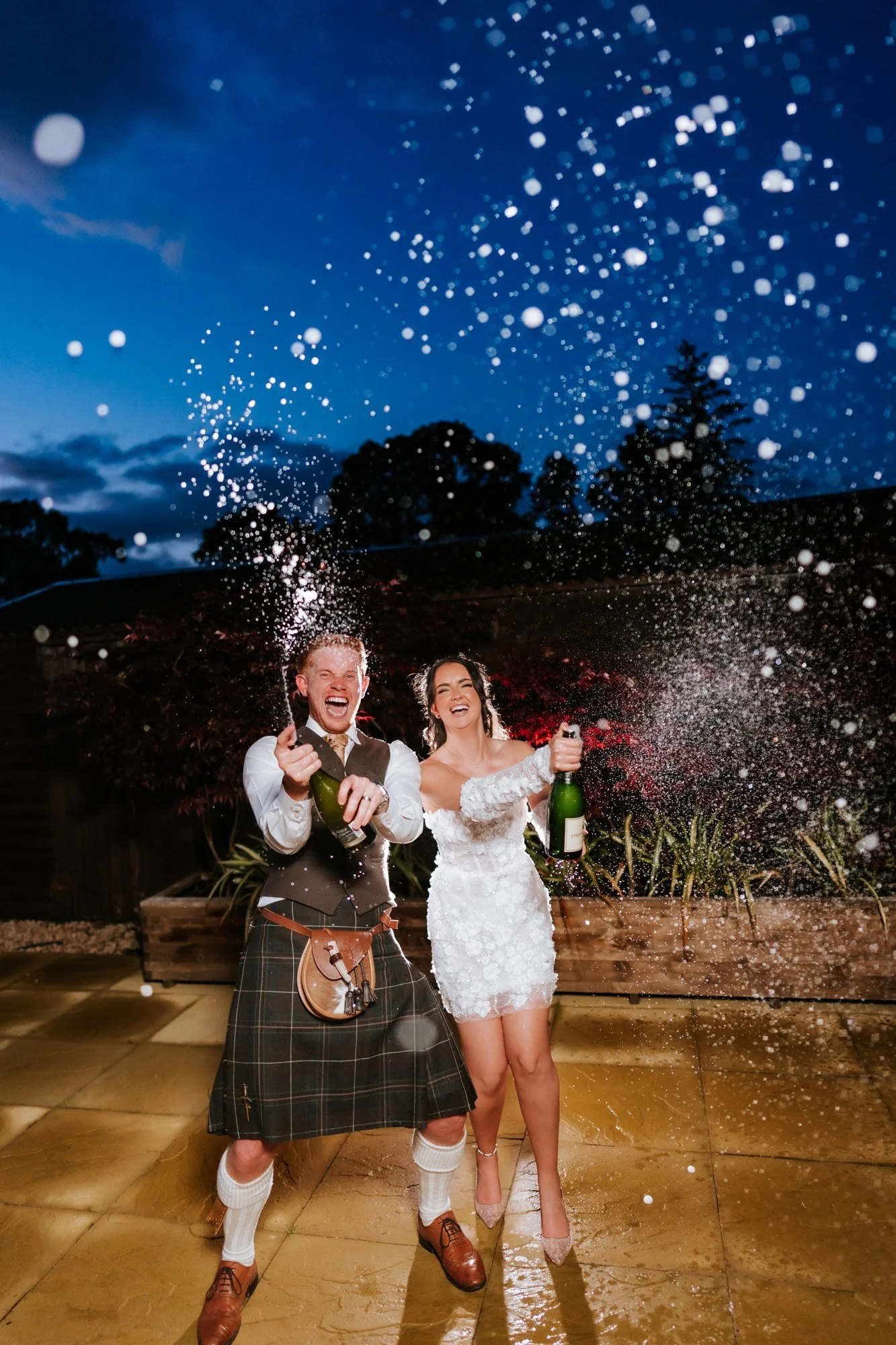 Kaitlyn and Scott spraying two champagne bottles beneath the evening sky at Glenbervie House.