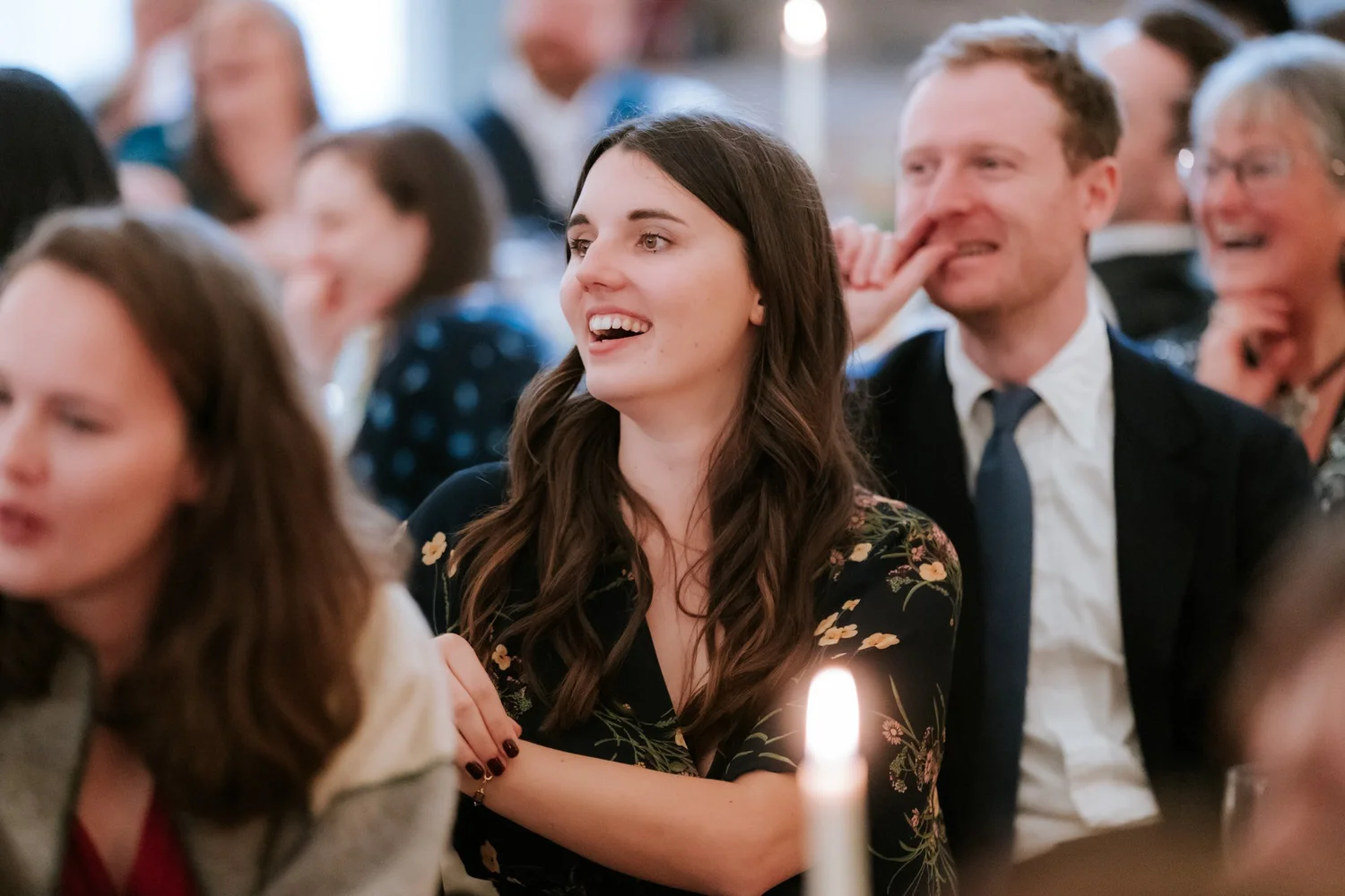 A guest in a floral dress laughs during the speeches, with smiling guests seated behind her.