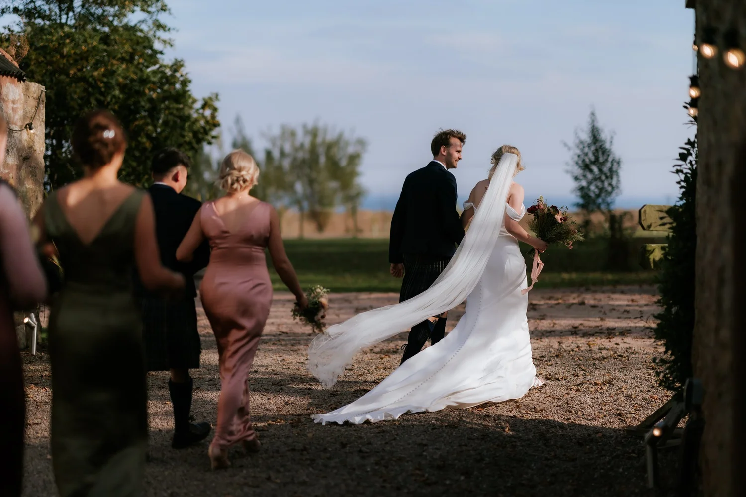 The couple walk past their guests after the outdoor ceremony, the bride’s veil trailing behind her.