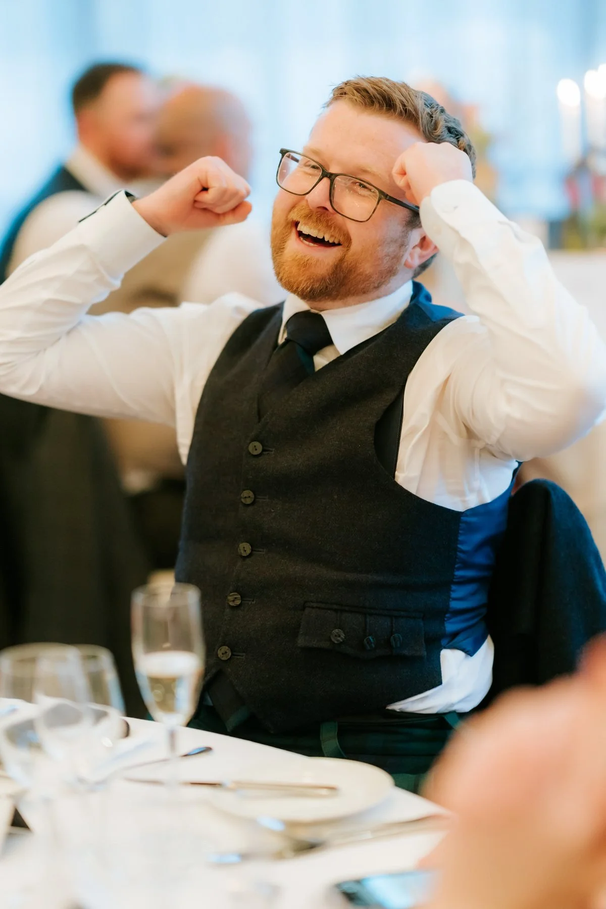 A guest in a black waistcoat laughing with his hands beside his head during the reception.