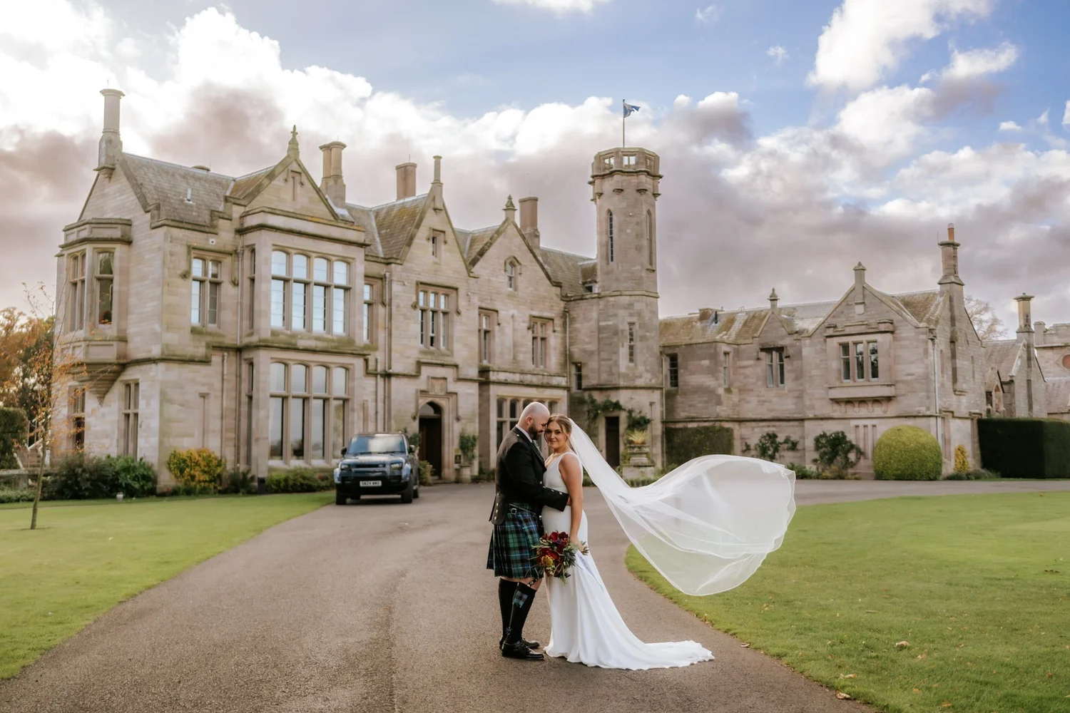 Kendall and Ewen standing together as her veil blows across the drive in front of SCHLOSS Roxburghe.