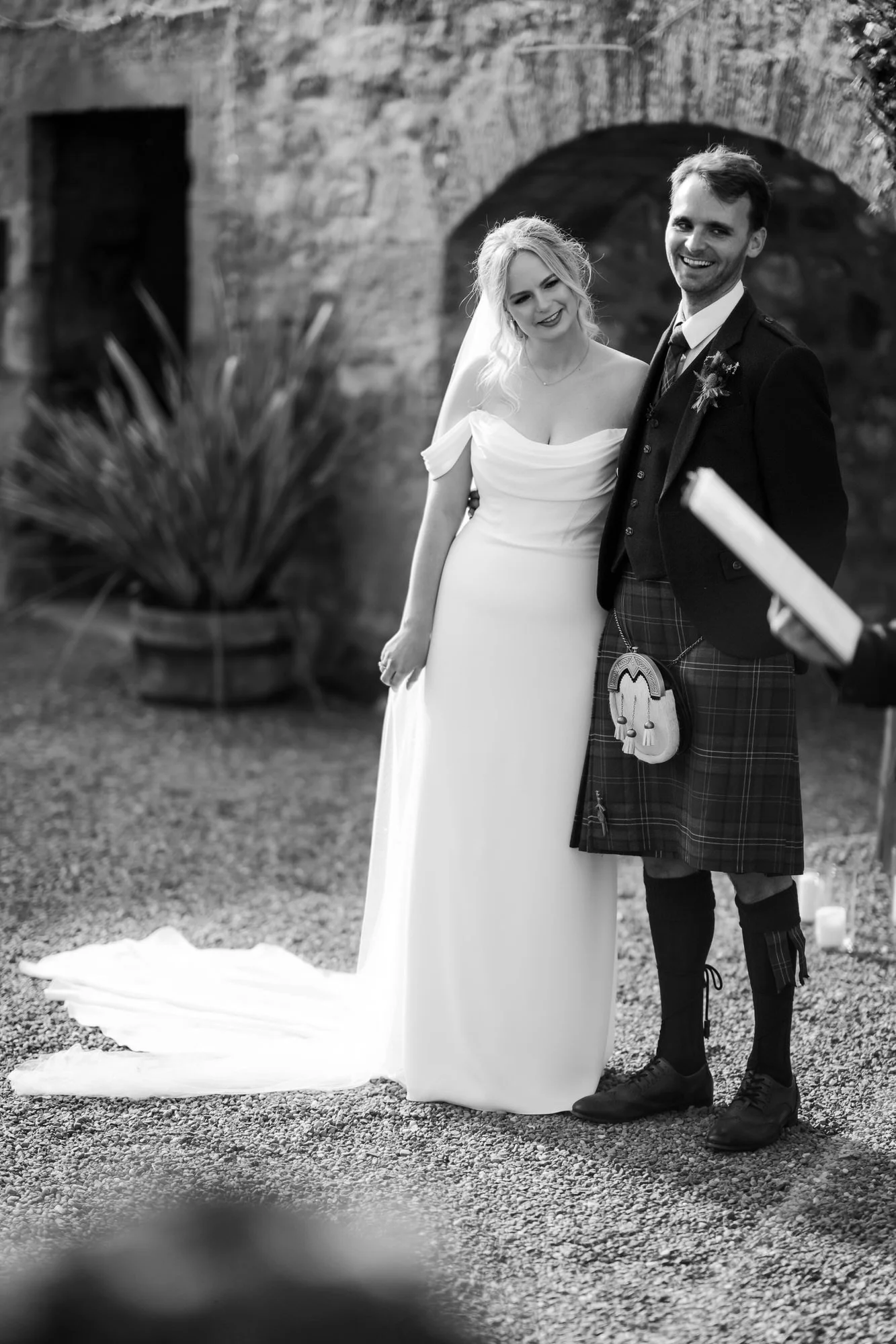 Black-and-white photograph of the couple standing arm in arm beside the stone arch during the ceremony.