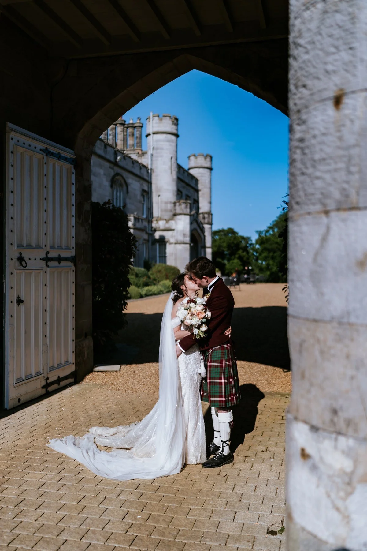Kim and Kenneth sharing a kiss beneath a shaded stone archway, with the castle beyond.