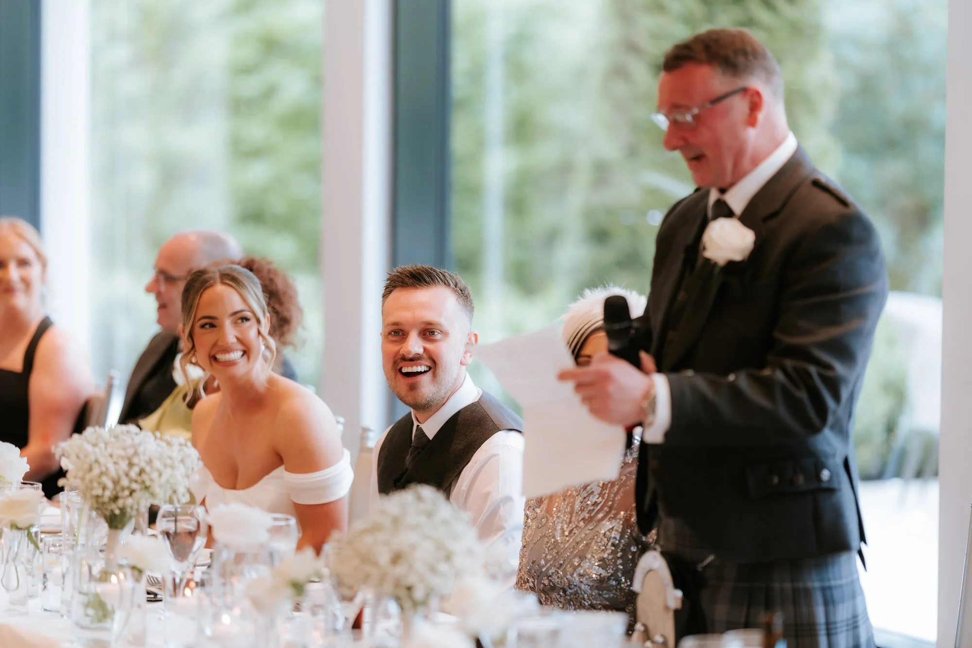 Elle and Adam laughing while a wedding speech is read at the top table