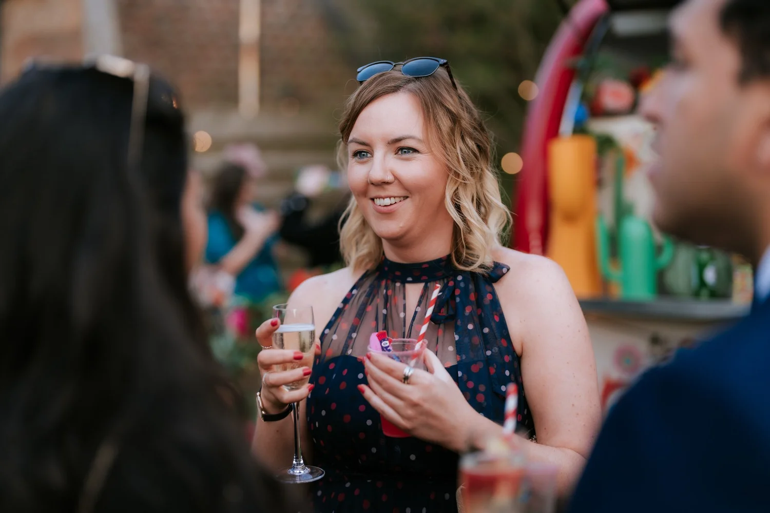 A guest smiles while holding two drinks during the outdoor wedding reception.