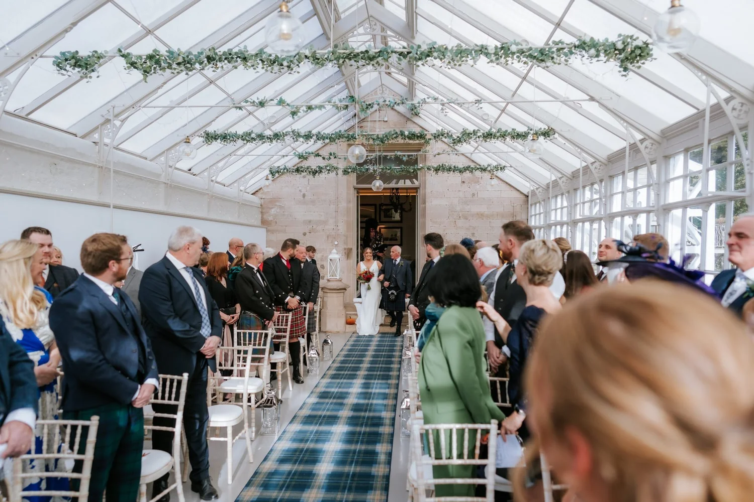 Guests standing as Kendall enters the bright conservatory for the wedding ceremony.