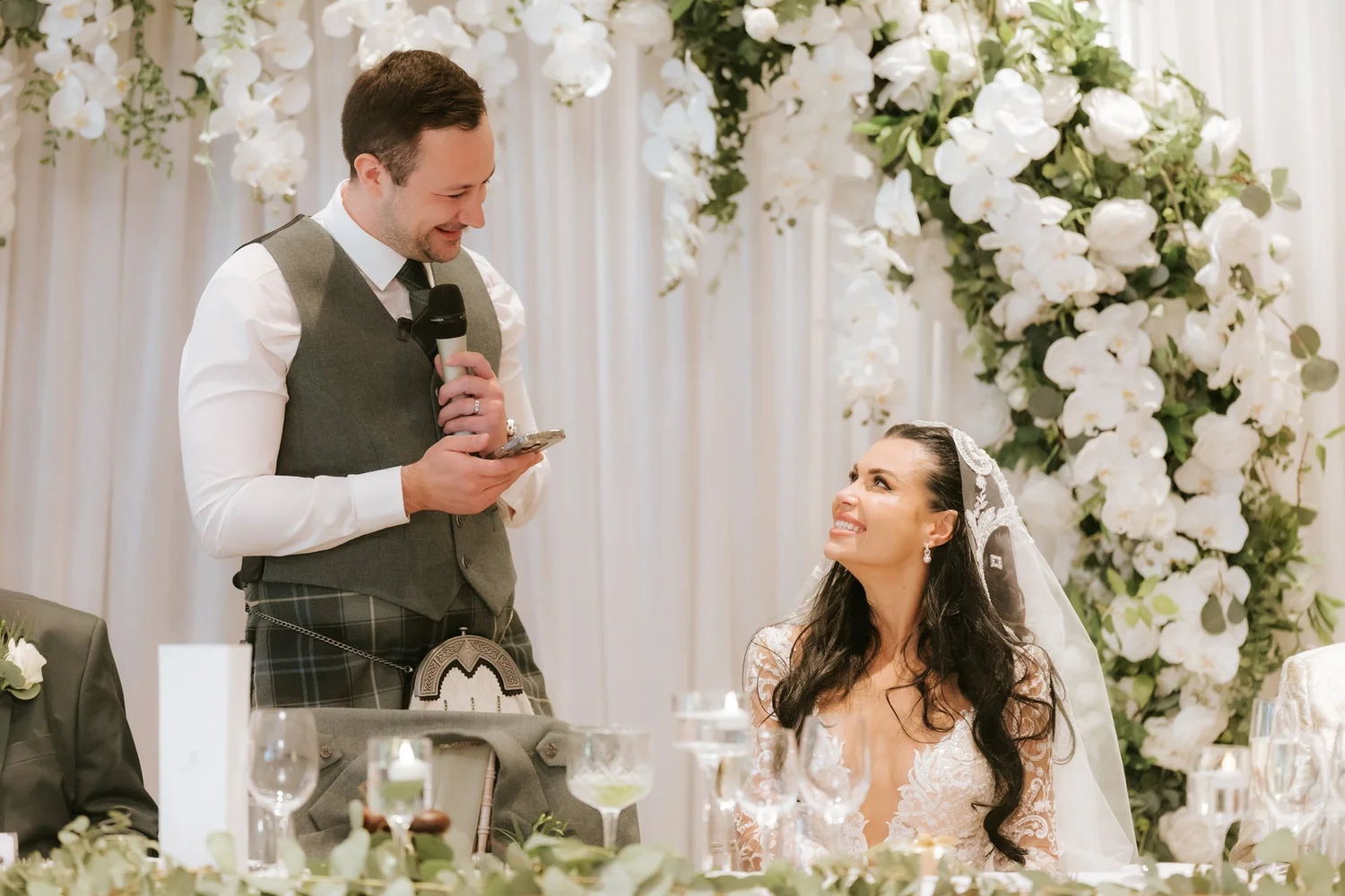 Connor speaking into a microphone at the top table while Cristina smiles up at him.