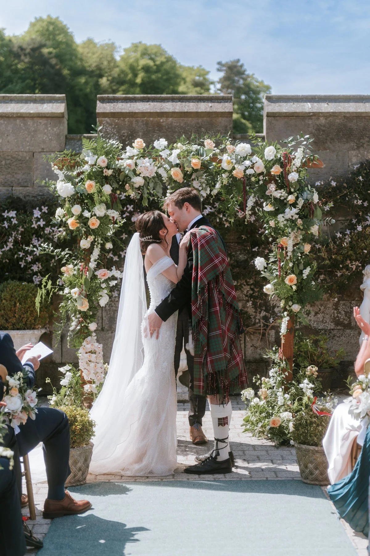 Kim and Kenneth sharing a kiss beneath the flower arch during their outdoor courtyard ceremony at Dundas Castle.