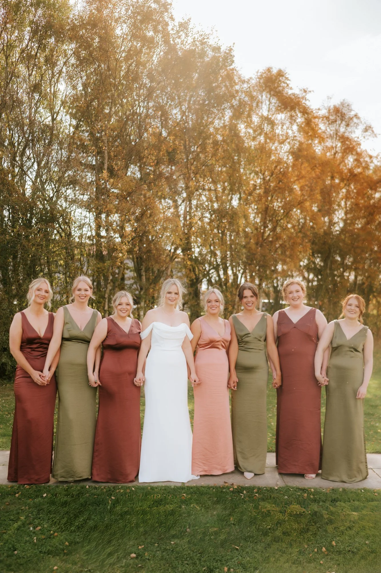 The bride stands with her bridesmaids in pink and green dresses on the lawn beside a tree.