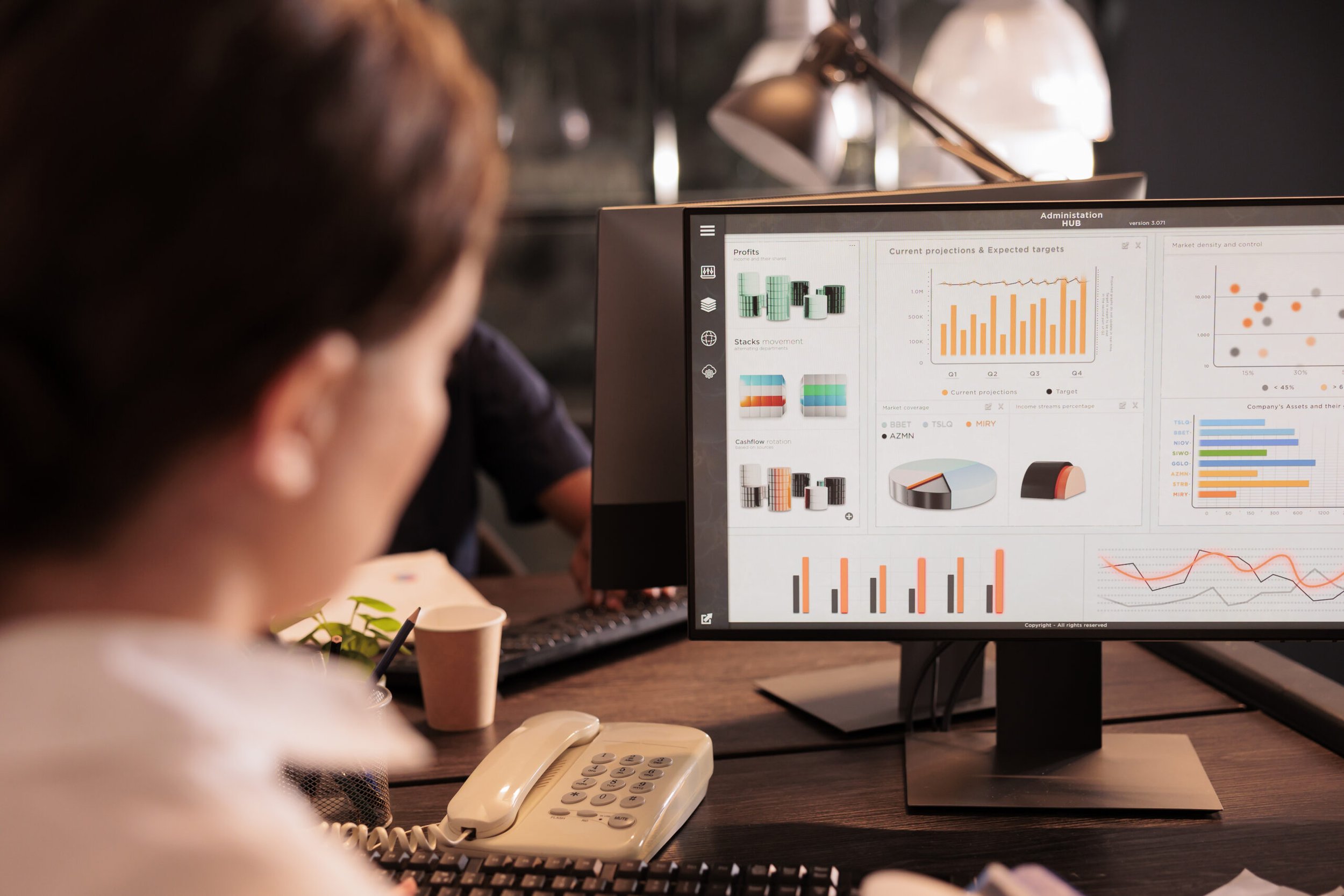 An analyst at a desk looks at a monitor showing a business dashboard with sales charts