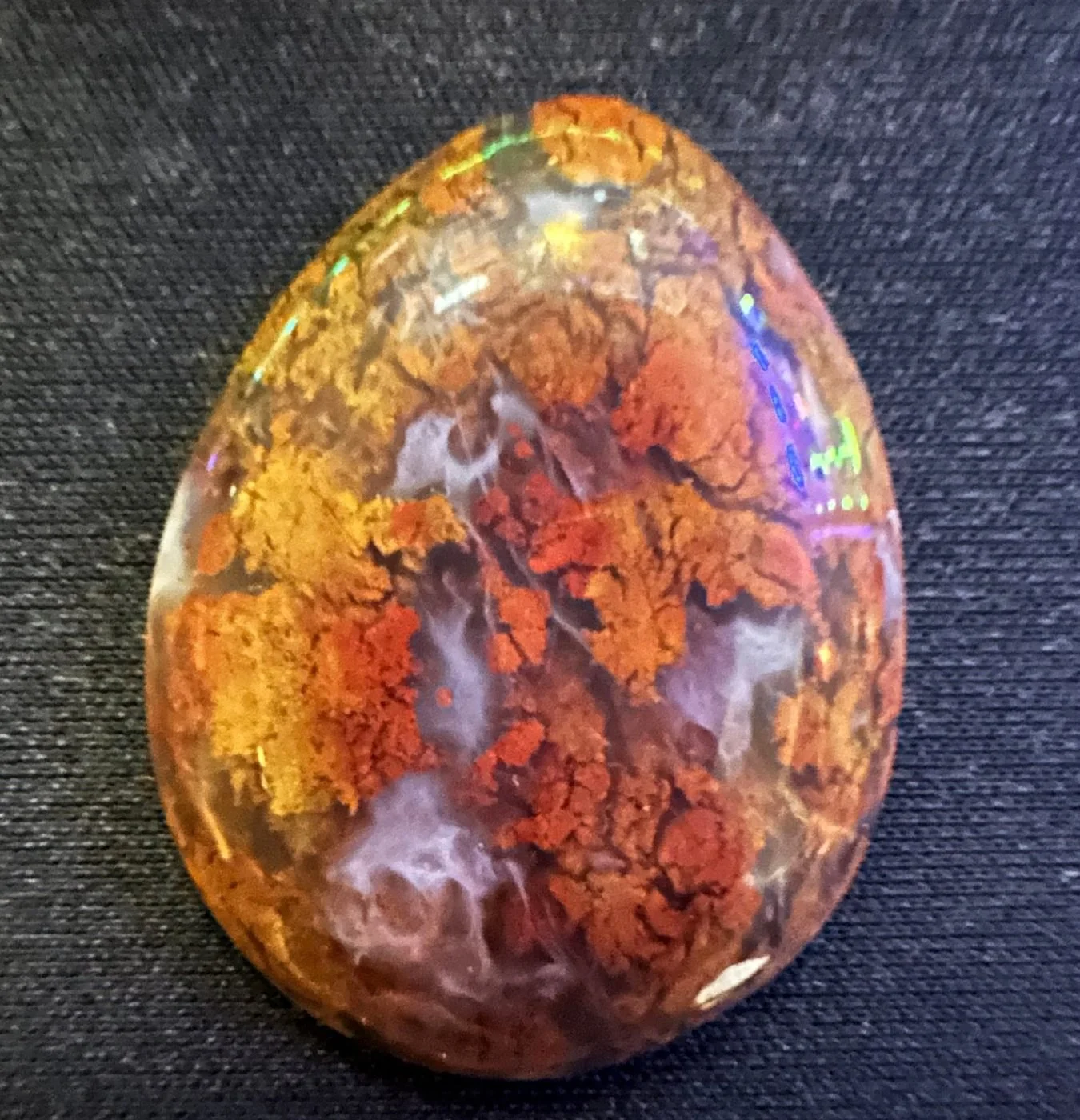 A polished cabochon of red, orange and gold jasper with milky chalcedony and flashes of rainbow color