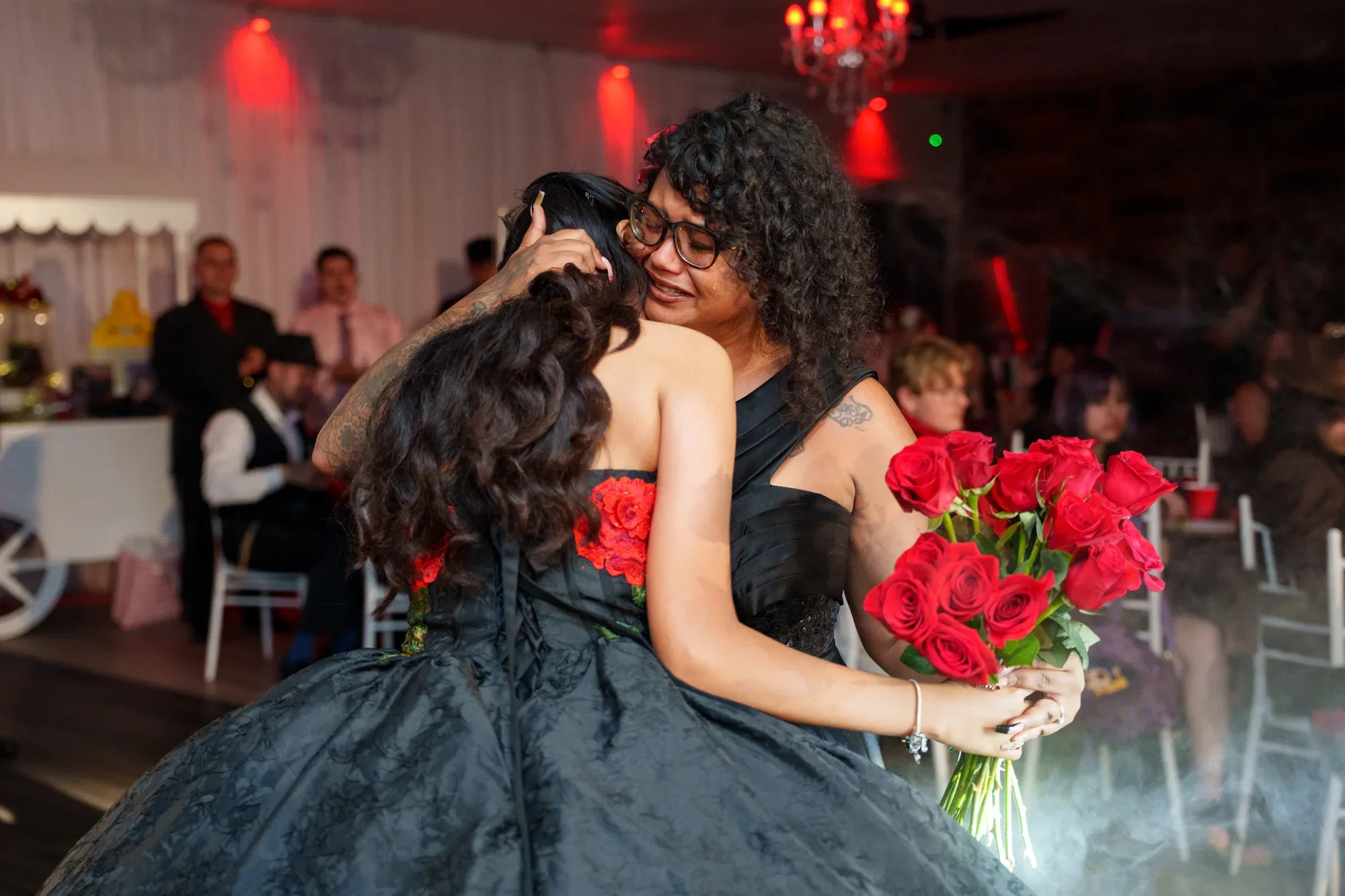 Una quinceañera abraza a su mamá durante su baile, con un ramo de rosas rojas en la mano