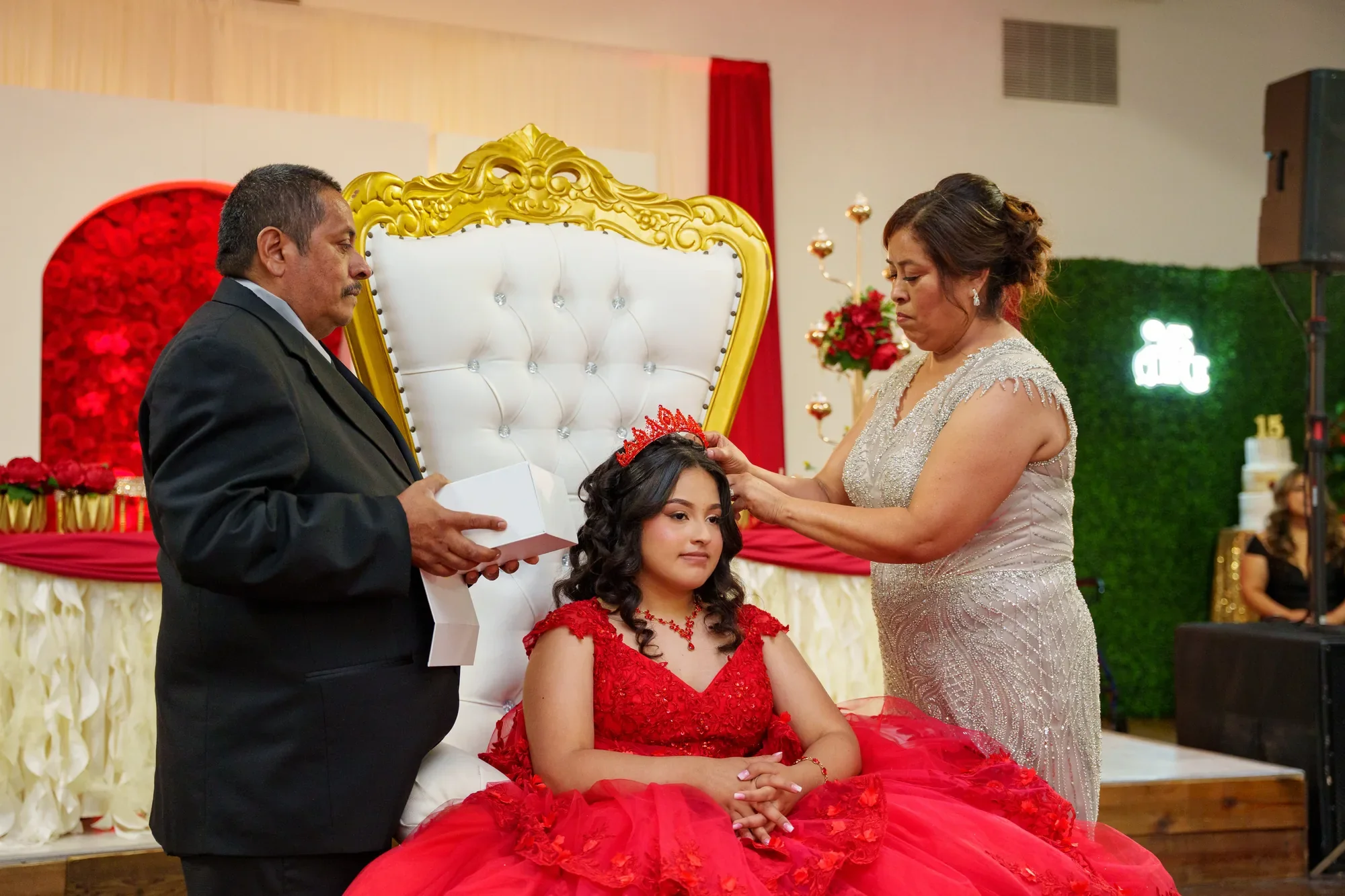 Los papás colocan la tiara a la quinceañera sentada en su silla real con vestido rojo