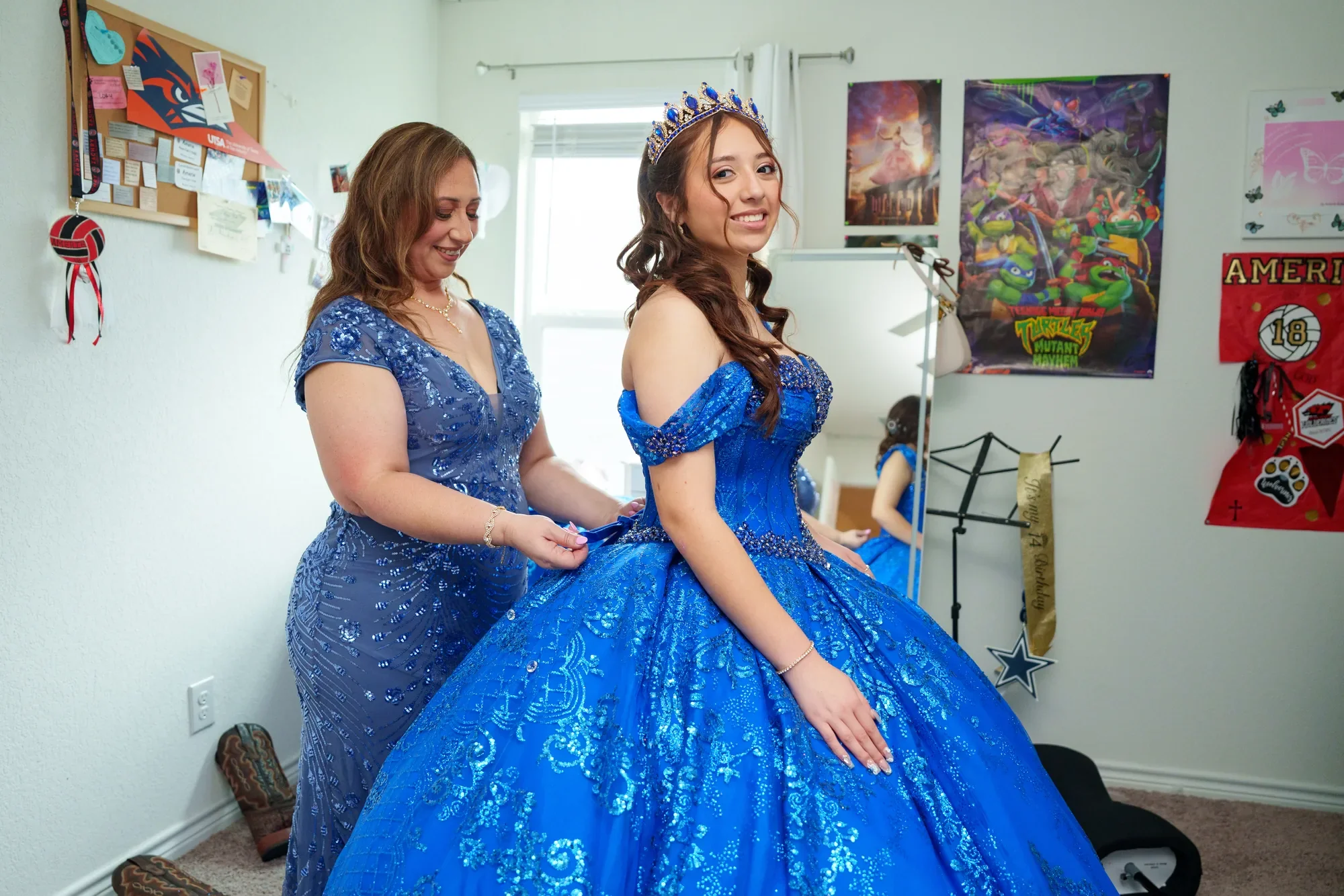 Mamá abrocha el vestido azul rey de la quinceañera en su habitación antes de la celebración