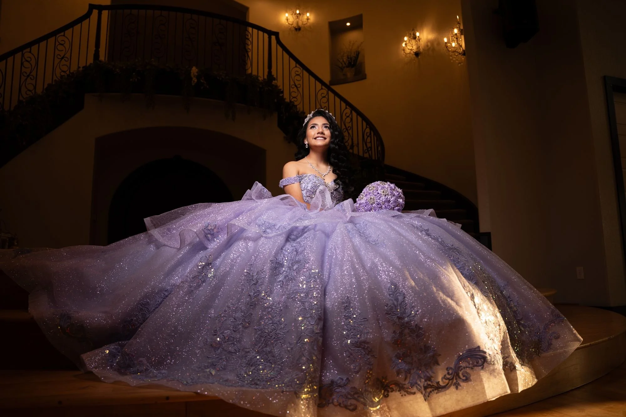 Quinceañera con vestido de bola lila brillante en una escalera imponente para una sesión de princesa de cuento de hadas