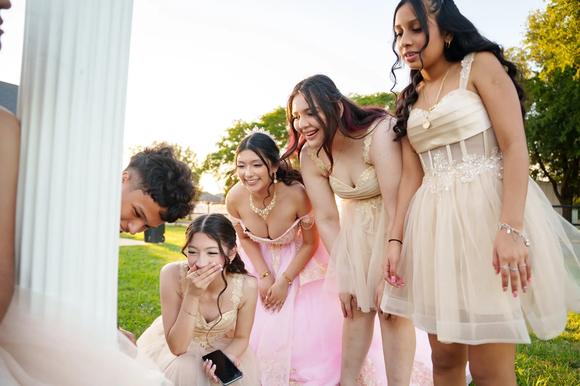 Una quinceañera y su corte se juntan alrededor de un celular riéndose al atardecer, planeando recrear un TikTok