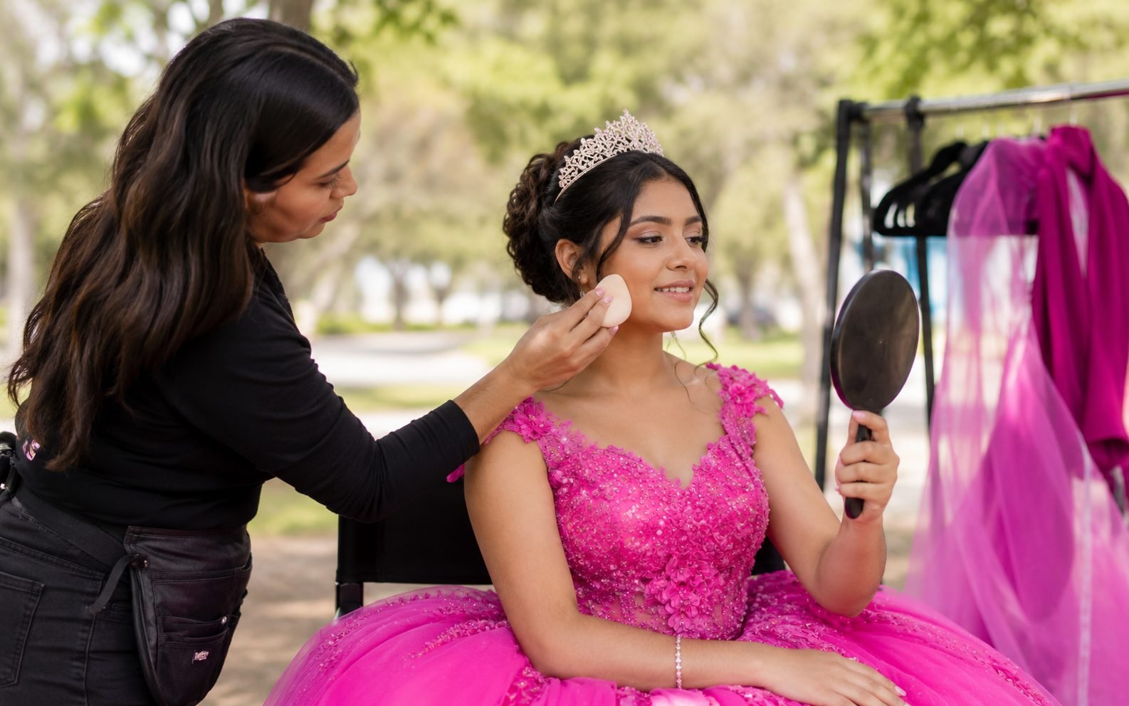 Estilista retocando el maquillaje de la quinceañera entre cambios de outfit mientras ella se mira en un espejo de mano con su corona
