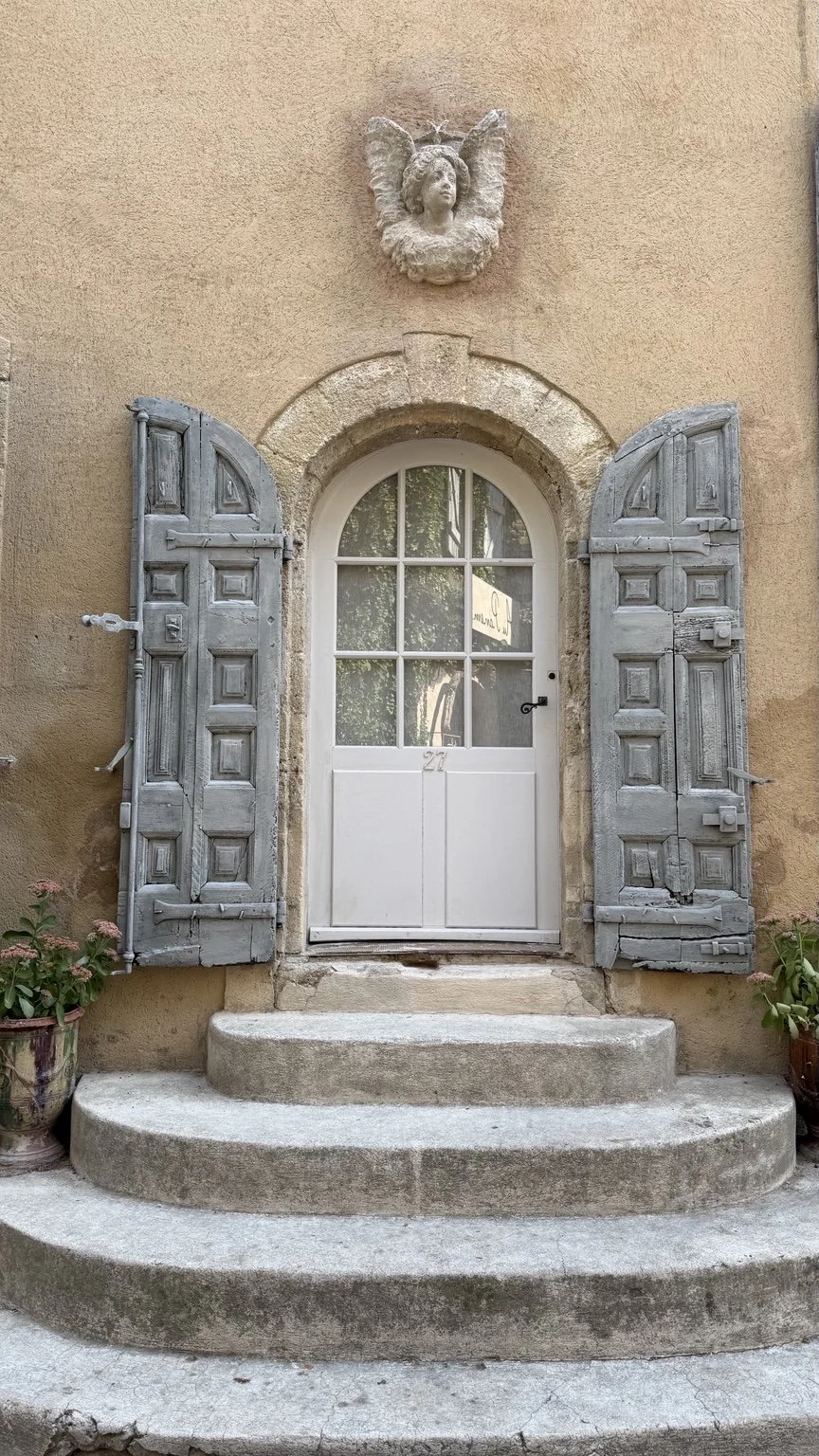 An arched white door with weathered blue shutters, stone steps and a carved angel above.
