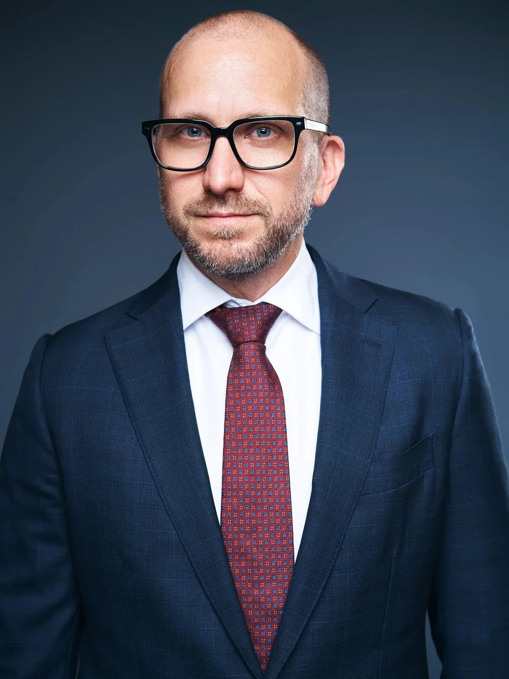 Close studio headshot of a lawyer in glasses, a navy suit and a red patterned tie
