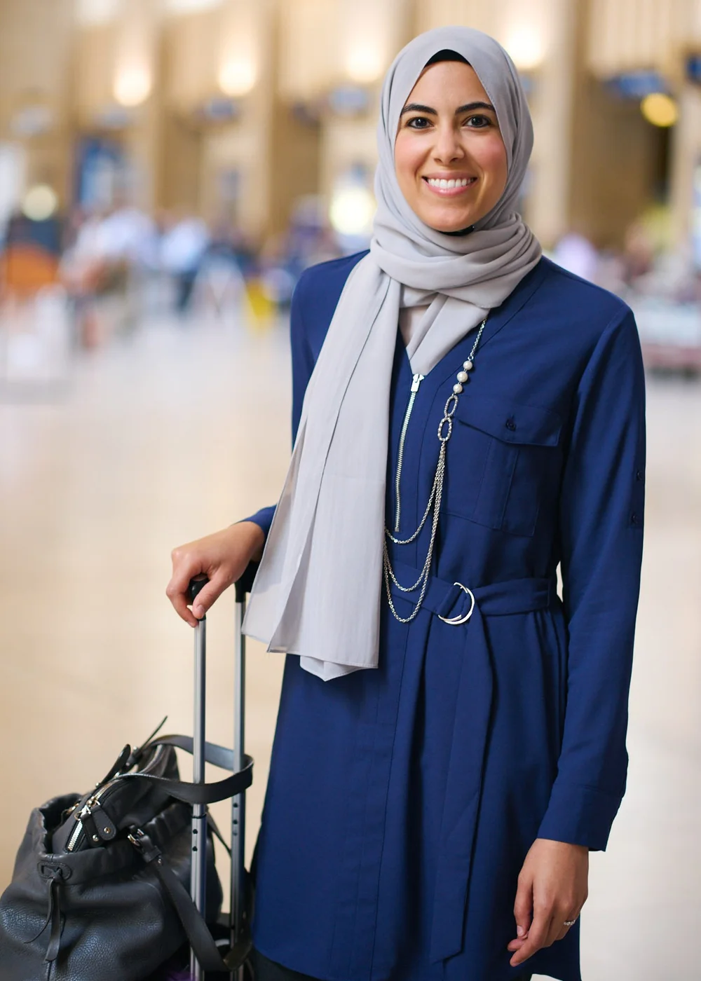 Travel portrait of a woman in a navy coat and light gray headscarf standing with luggage in a railway concourse
