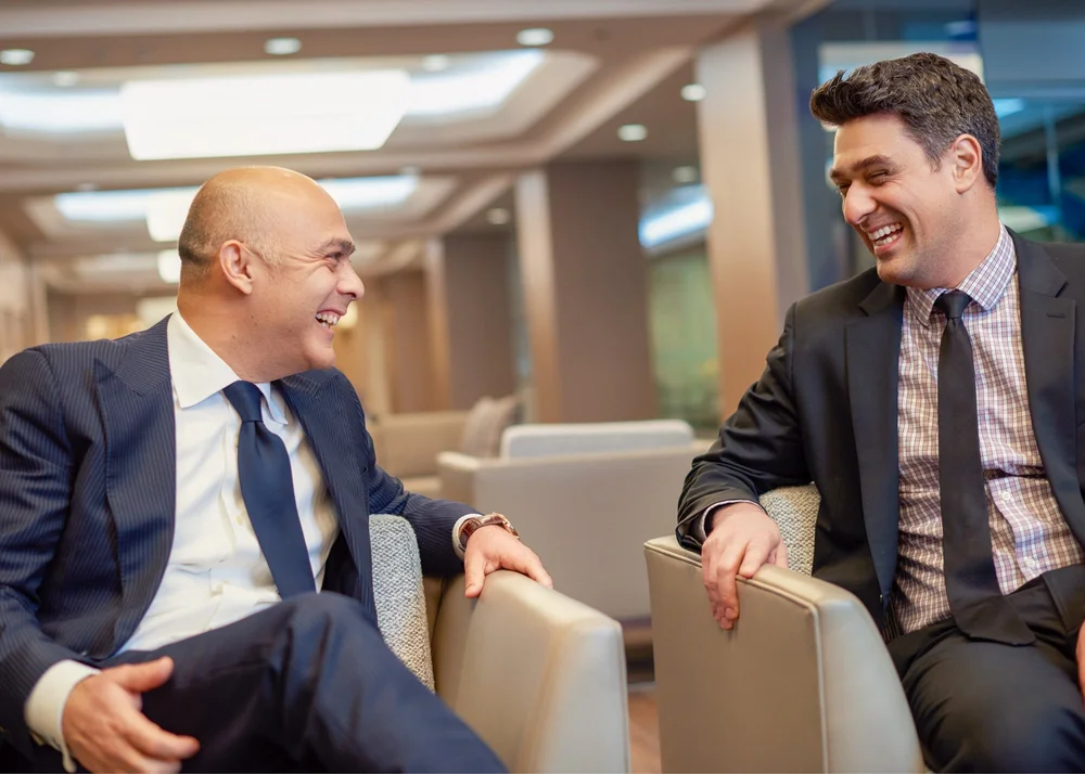 Two colleagues in suits laughing together in a corporate office lounge